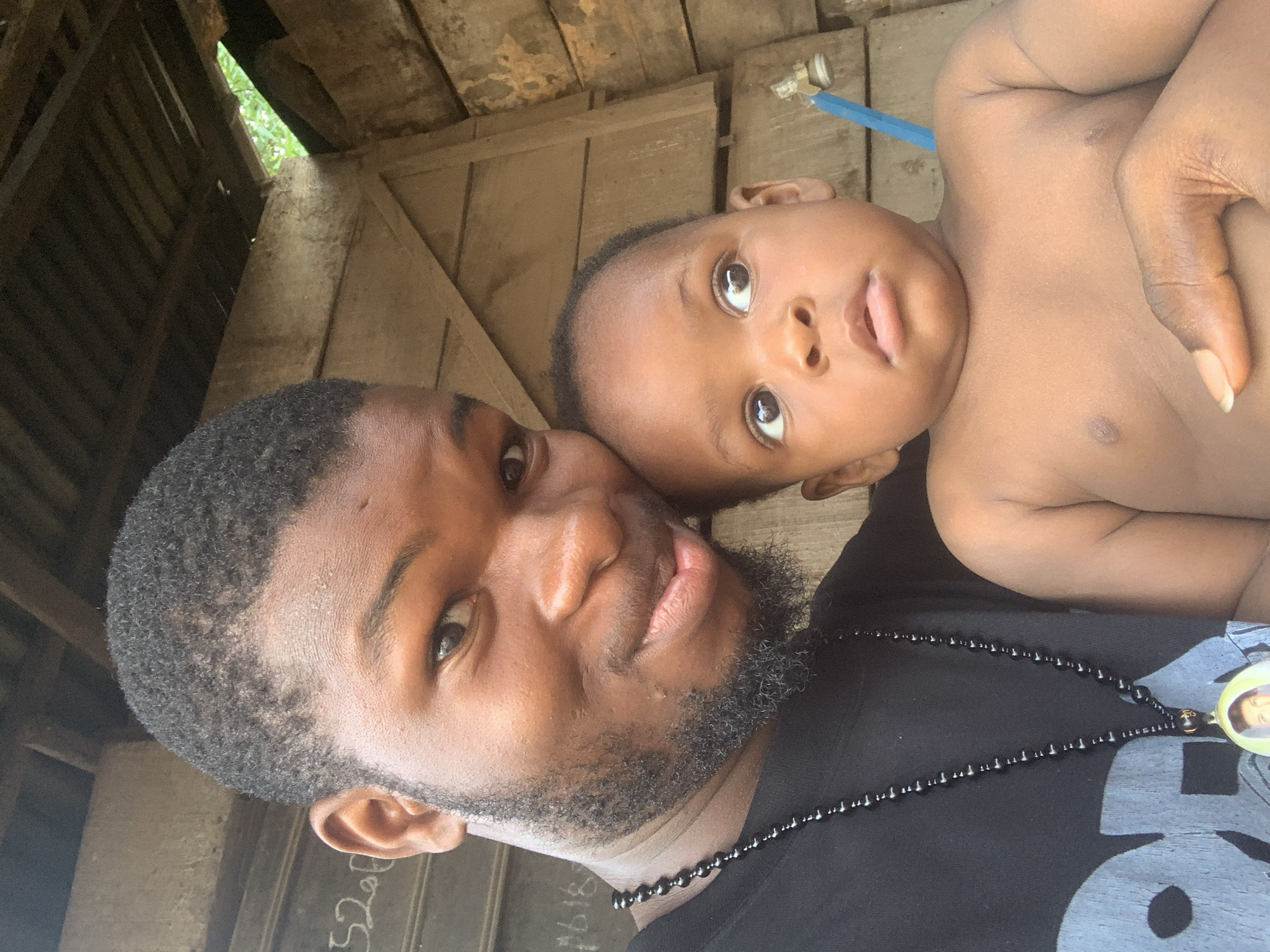 The image displays a man and a baby in a close-up, selfie-style portrait within a rustic indoor or sheltered setting, likely in Nkpor, Nigeria.
The man, positioned in the foreground and to the left, has dark skin, short dark hair, and a neatly trimmed beard. He is wearing a black t-shirt and a long black beaded necklace with a circular pendant featuring a faded image, possibly a face. He looks towards the camera with a gentle, slight smile.
Cradled by or positioned closely next to the man is an infant with dark skin, large wide-open eyes, and a bald head. The baby appears to be shirtless or wearing a very light-colored top, and looks upwards with a curious expression and a slightly open mouth. A hand, presumably the man's, is visible holding the baby.
The background is characterized by rough, untreated wooden planks, suggesting an unsophisticated structure such as a shed, shack, or an informal dwelling. Gaps in the wooden construction allow bright daylight to illuminate the scene and reveal lush green foliage outside, indicating an outdoor element nearby. A small blue pipe or hose fixture is visible in the upper right portion of the background. On a wooden plank behind the man, some handwritten white text and numbers are visible, including "352," "2020," and "2016."
The lighting suggests it is daytime. The overall impression is one of warmth and affection between the man and the baby in a simple, natural environment.