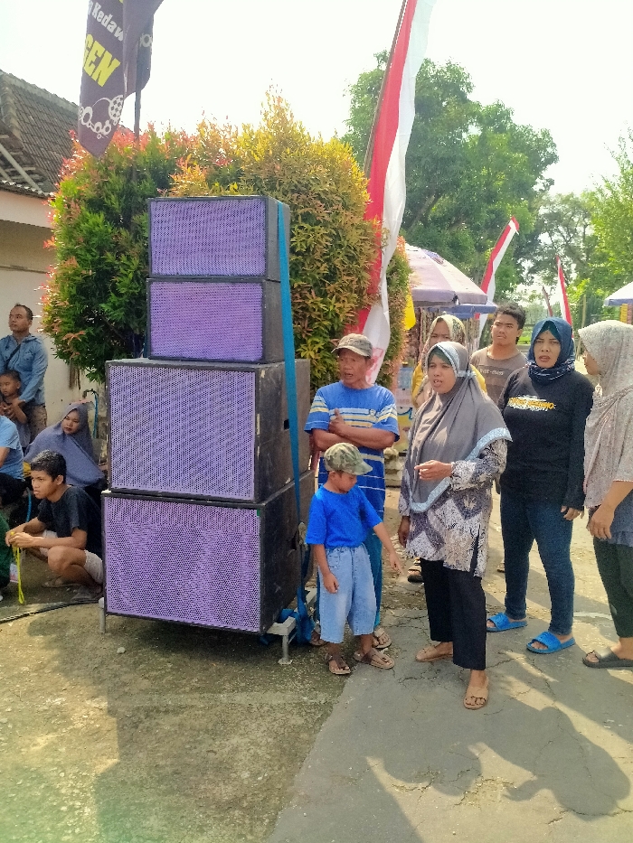 The scene is located in Kecamatan Kedawung, Indonesia, at the coordinates latitude -7.514, longitude 111.03. There is a stack of four large speakers with purple grilles on a paved area in front of a group of people. The speakers are secured with blue straps. A young boy wearing a blue shirt and green cap stands next to the speakers. To his right is a man in a blue and white striped shirt, with his arms folded across his chest. A woman wearing a gray headscarf and patterned dress stands to the right of the man, her hands clasped in front of her. A woman wearing a black shirt with a white emblem on the front and black pants stands to the right of the woman in the gray headscarf. A woman wearing a white headscarf with a black dress stands to the right of the woman in the black shirt. A man wearing a red shirt and khaki pants stands to the right of the woman in the white headscarf. Two children stand in the background, one on each side of the speaker stack. A man in a dark blue shirt stands behind the children. In the background, there is a red and white flag, greenery, and a white tent.