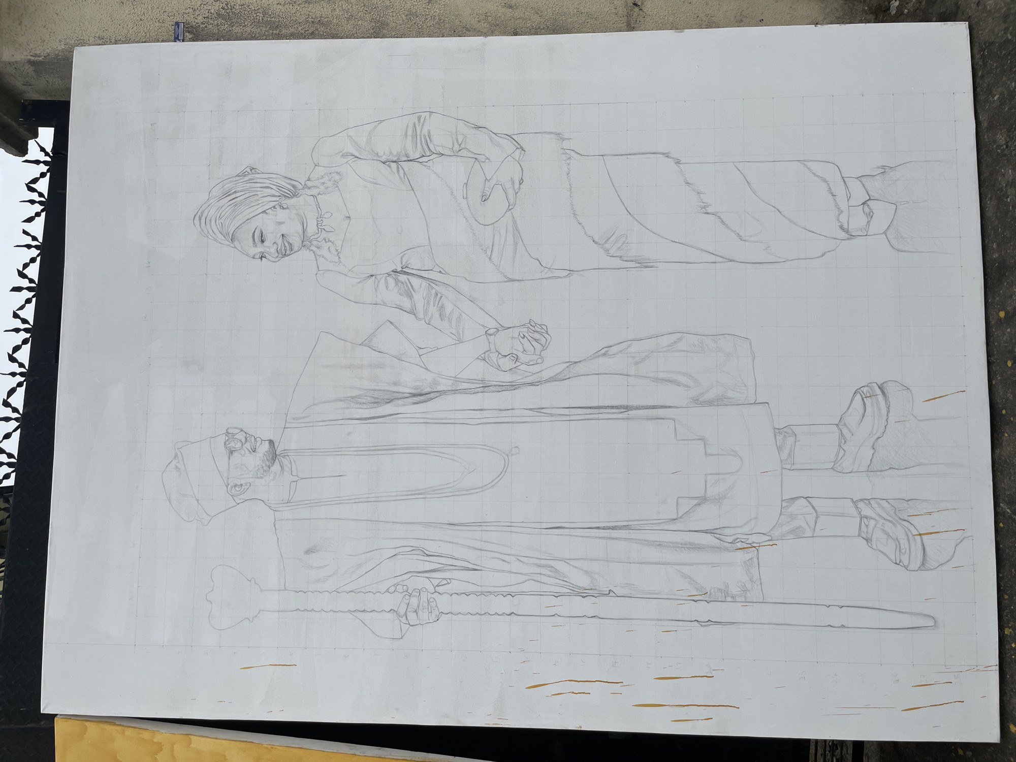 A large, rectangular white canvas is laid horizontally on a likely outdoor surface in Ado, Nigeria. A pencil drawing of two figures, a man and a woman, is sketched on the canvas. A grid is lightly drawn in pencil across the entire surface, dividing the drawing into squares.
The man is drawn in the lower portion of the canvas. He is bearded, wearing a draped garment with a rectangular decoration on the chest and ornate shoes. He holds a long, carved staff.
The woman is drawn above the man, tilted slightly. She also wears draped clothing and a head wrap. Her hands are clasped at her waist.
The canvas shows signs of paint splatter in yellow/brown color. The edges and corners are imperfect. A black metallic ornamental grate sits to the left. The background consists of a overcast sky and a concrete surface.