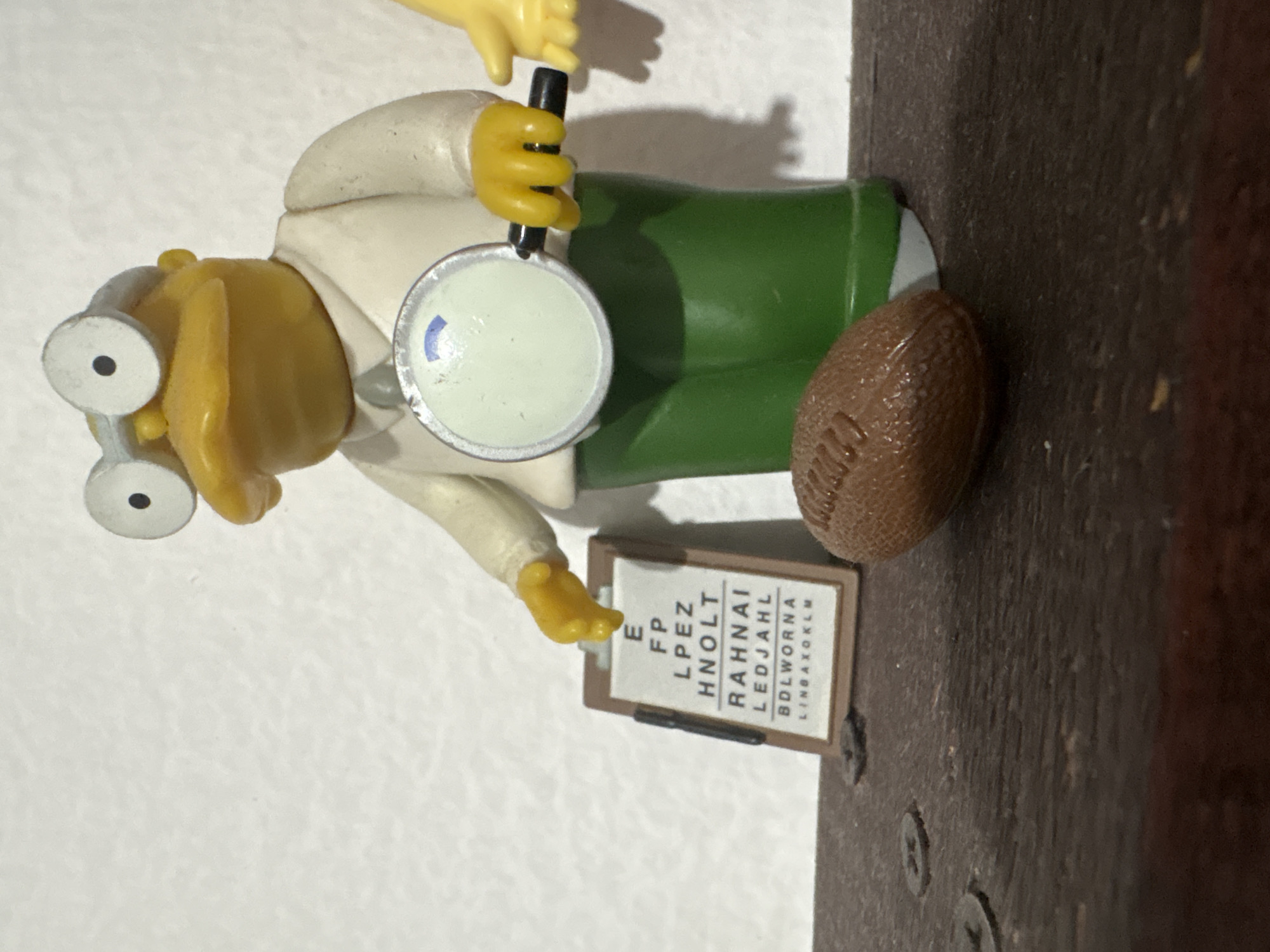 An eye chart and a football are positioned in front of a yellow, cartoon-like figurine wearing glasses, a white shirt, and green pants, holding a magnifying glass. The figurine appears to be an optometrist or doctor. The scene is set on a dark, wooden surface against a white background. The image is a close-up shot, suggesting it is an indoor scene. There is no indication of the city of Cuernavaca, Mexico, or any specific urban environment.