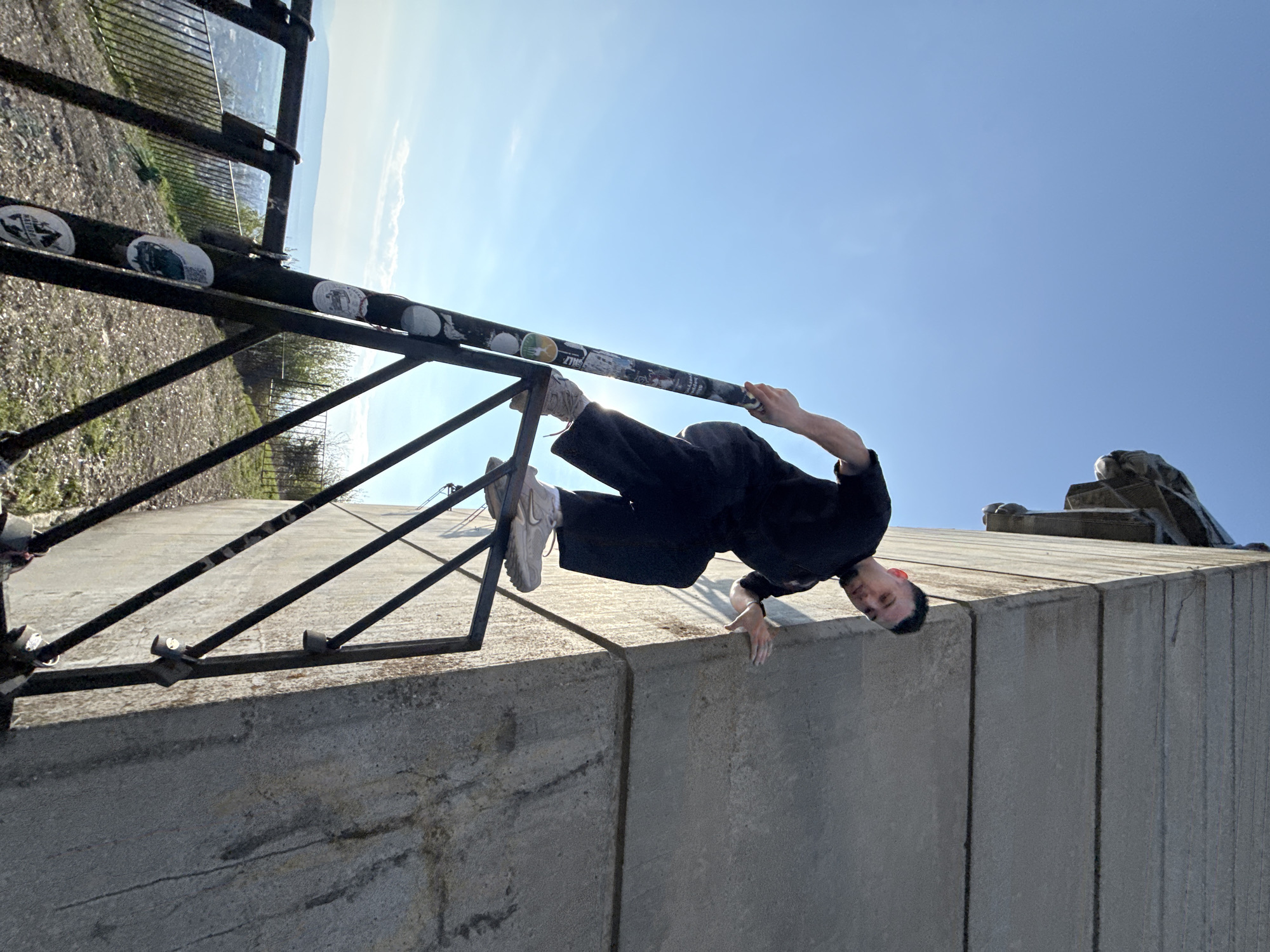 A person in dark clothing and light-colored sneakers is hanging upside down from a black metal railing affixed to a light gray concrete structure. The railing is covered with stickers. The person is grasping the railing with one hand and touching the concrete structure with the other. In the background, there is a view of the city. The location is likely the monument "Founders of the Bulgarian State" in Shumen, Bulgaria.