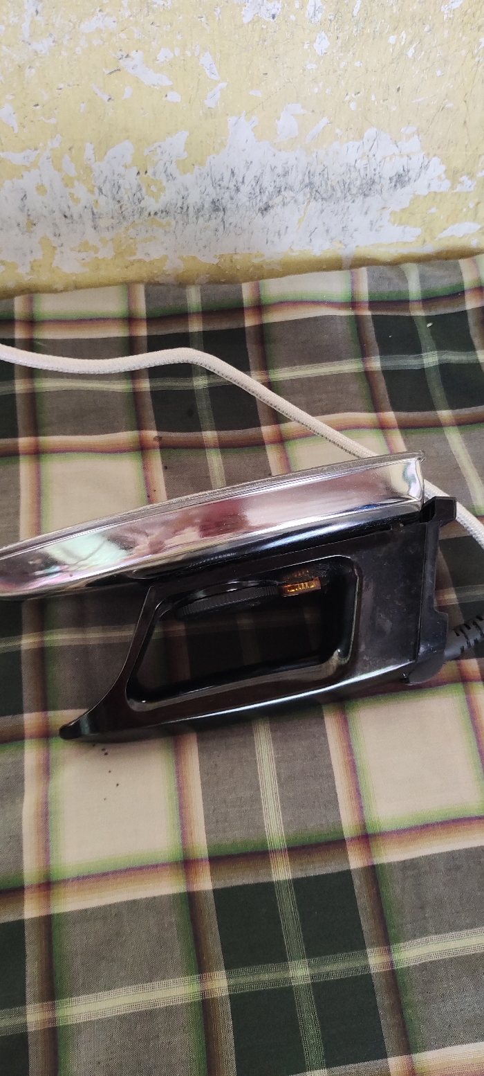 The image shows a black and silver electric iron resting on a green and white checkered fabric. The iron is plugged in, with a white electrical cord visible. The iron is sitting on a surface that appears to be a table or bed. The iron is in the foreground of the image, with the fabric taking up the background. The cord of the iron is visible at the bottom right corner of the image. The iron is a typical household appliance used for pressing clothes. The coordinates provided (latitude: -7.828, longitude: 112.701) correspond to a location in Kecamatan Lawang, Indonesia. However, there is no way to determine if the image was actually taken at that location. The image is likely taken inside a residential home.