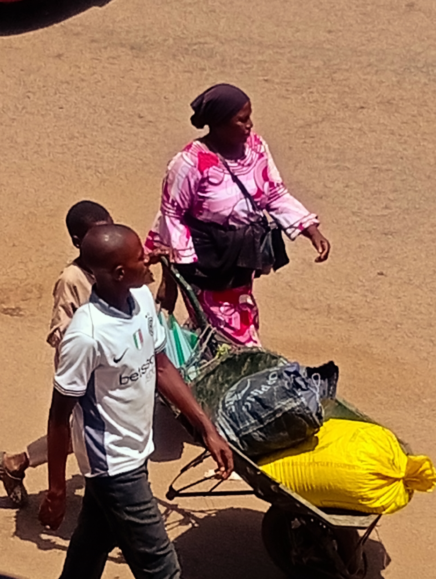A group of people are seen walking on a dirt road in Bukuru, Nigeria. In the foreground, a young man in a white t-shirt and dark pants is pushing a wheelbarrow. The wheelbarrow is loaded with two large bags, one dark and one bright yellow. Behind the young man, a woman in a pink and white patterned dress and a dark headscarf is walking. Partially obscured behind her and to her left, a young boy in a light brown shirt is walking. The ground is dusty and uneven, suggesting a rural or unpaved urban area. Shadows suggest the sun is high in the sky.