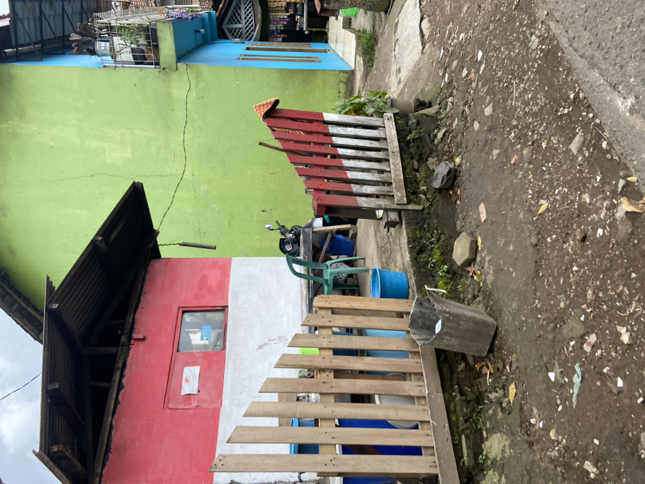 The image shows a view of a street in Lembang, Indonesia, located at coordinates -6.835 latitude and 107.602 longitude. Two adjacent houses are visible, one painted in red, white, and green with a black roof, and the other in light green and blue, with a white roof. Both have wooden fences in front. There is a motorcycle parked outside the red, white, and green house. A wooden fence in front of the red, white, and green house has fallen over, and is propped against the wall. A blue bucket is visible on the ground. The ground is covered with dirt and debris.