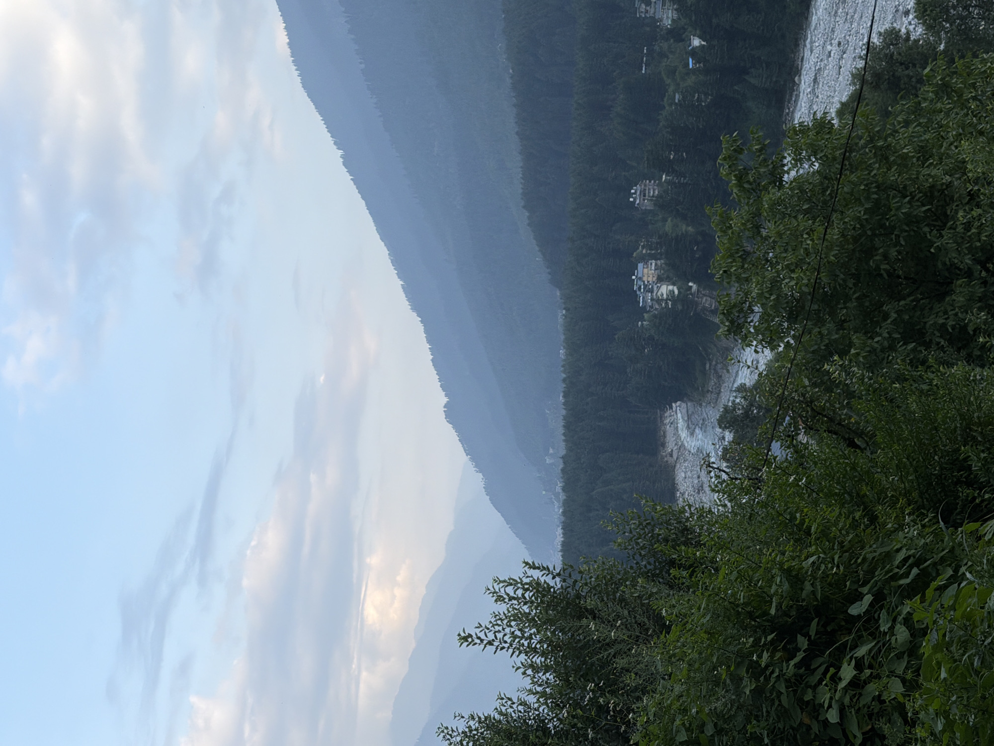 The image depicts a panoramic view of a valley with rolling hills and a river. In the foreground, lush green foliage frames the scene. The middle ground is dominated by a dense forest, followed by a winding river with a rocky bed. Several buildings, possibly hotels or residences, are scattered amidst the trees along the riverbank. The background features layers of misty mountains fading into the hazy sky, suggesting a vast, natural landscape. The sky above is a soft blue with scattered clouds, hinting at either early morning or late evening light. The location is identified as Vashisht, India, a place known for its natural beauty and scenic views. There are no people visible in the image.