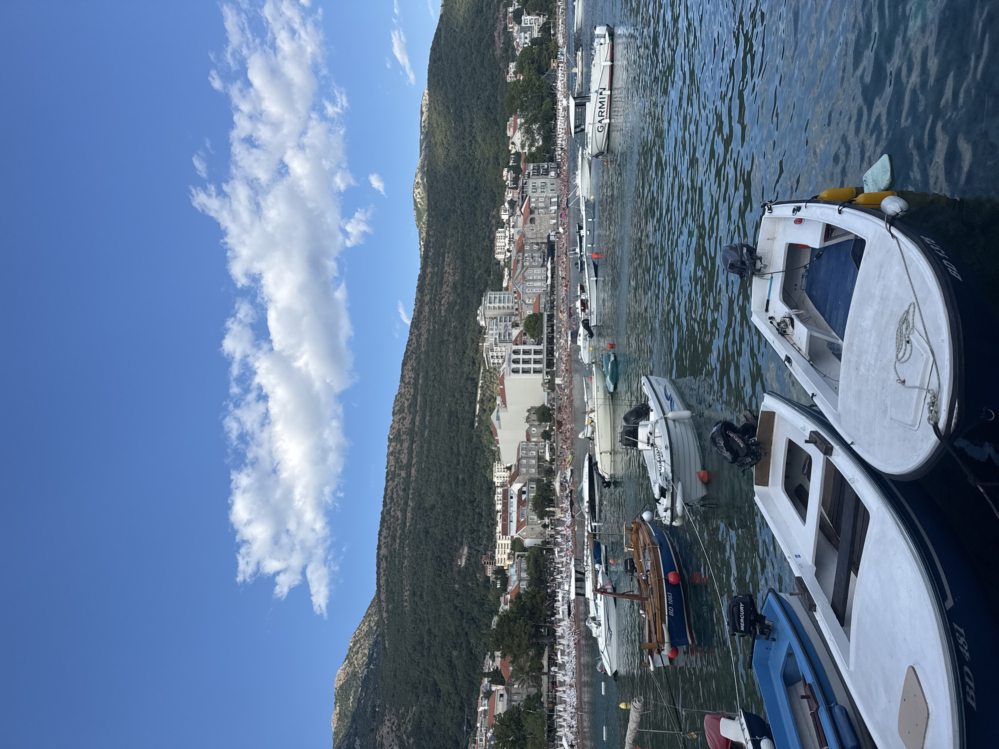 The image captures a sunny day in Petrovac centar, Montenegro, with a clear blue sky dotted with white cumulus clouds. The foreground is dominated by a calm, blue body of water filled with various boats. Several small motorboats are moored, some with outboard motors visible. One boat in the center foreground is white with blue trim and the registration "BD 481" is visible. To its right, another white boat with a blue interior and yellow buoys is also present. Further back, a classic wooden boat with the registration "BD 1063" is moored.
In the mid-ground, a bustling beach is lined with rows of white beach chairs and umbrellas, indicating a popular tourist destination. People are visible on the beach, enjoying the sun and sand. Behind the beach, a picturesque town unfolds, characterized by a mix of traditional stone buildings and modern multi-story structures. The town is nestled at the base of a lush, green mountain that rises steeply in the background, creating a dramatic natural backdrop. The overall scene depicts a vibrant coastal town on a pleasant day.