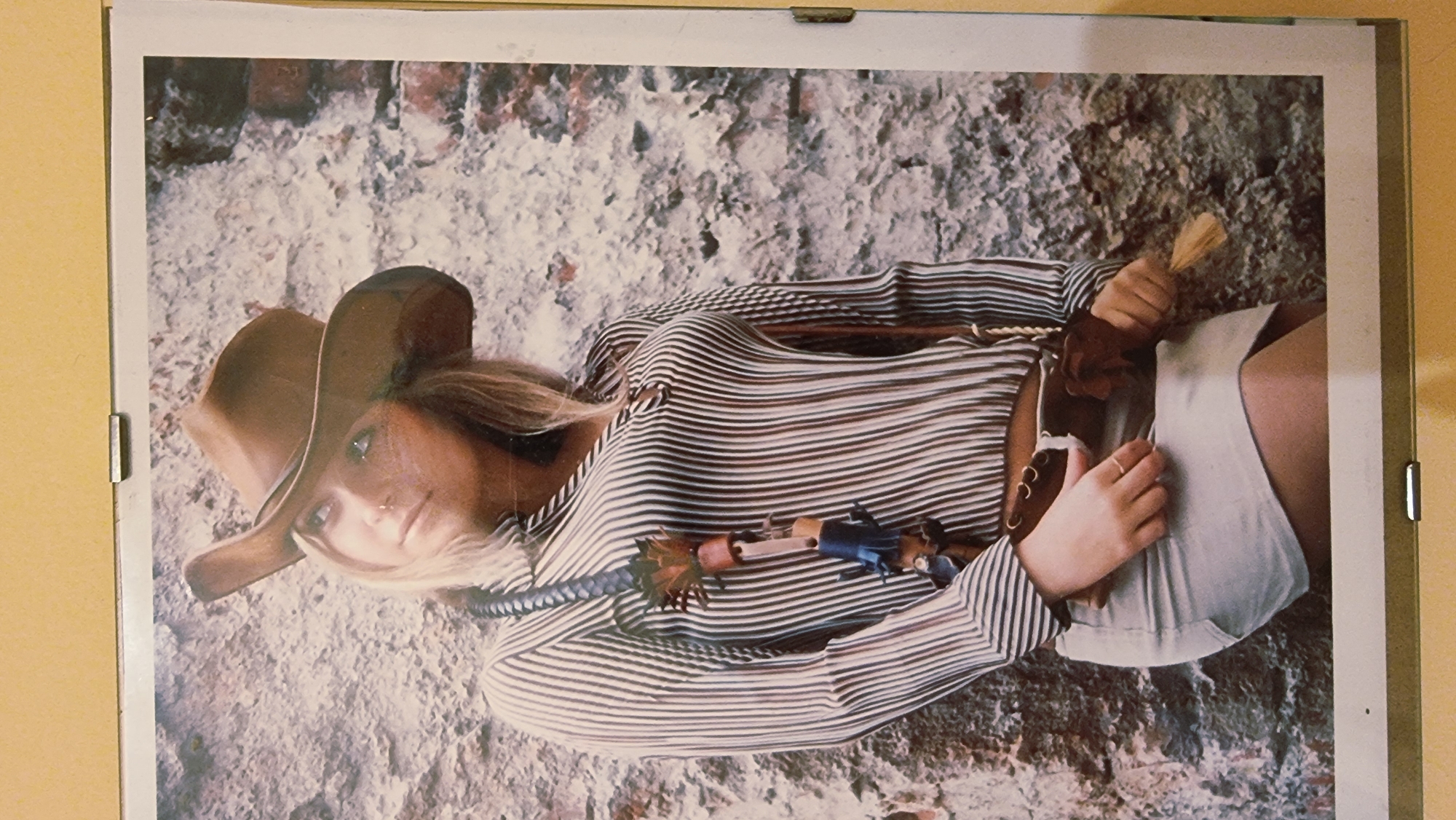 A woman, positioned centrally and angled slightly to the left, stands against a textured stone wall. She wears a brown cowboy hat, a vertically striped long-sleeved shirt, and a light-colored mini-skirt. A decorative strap with tassels hangs from her shoulder, and a brown belt with a holster is visible around her waist. Her left hand rests on the holster, and her right hand is near the holster's opening. The background is a roughly textured wall, suggestive of weathered stone or brick, with some darker, more intact brick sections on the right. The lighting appears to be natural, casting subtle shadows. The overall image is a photographic portrait. The location is not discernible from the image itself.