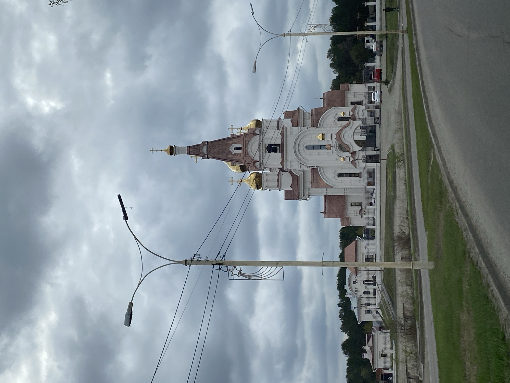 The image shows a church in Shelekhov, Russia. The church has a white exterior with brown accents and golden domes. It's located next to a road. Streetlights and power lines are visible in the foreground. The sky is overcast with gray clouds. The church appears to be situated in a residential area.