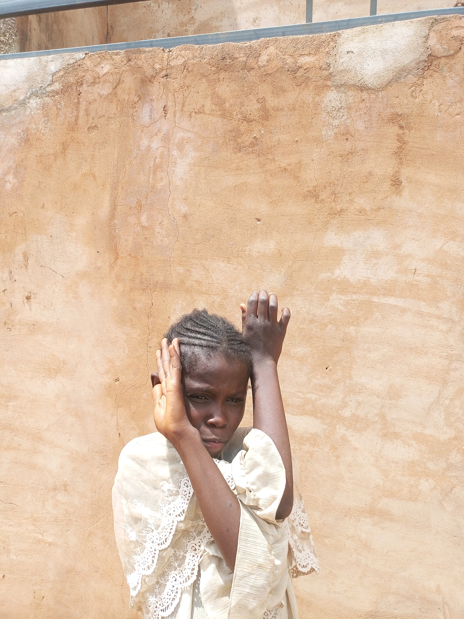 A young girl stands in front of a textured, light brown wall, her face partially obscured by her hands. She wears a light-colored dress with lace trim. The wall shows signs of wear and tear, with cracks and patches of lighter material. Above the wall, a metal railing is visible. The scene appears to be outdoors in an urban setting. The lighting suggests it is daytime. The girl's posture and expression suggest a moment of contemplation or shyness.