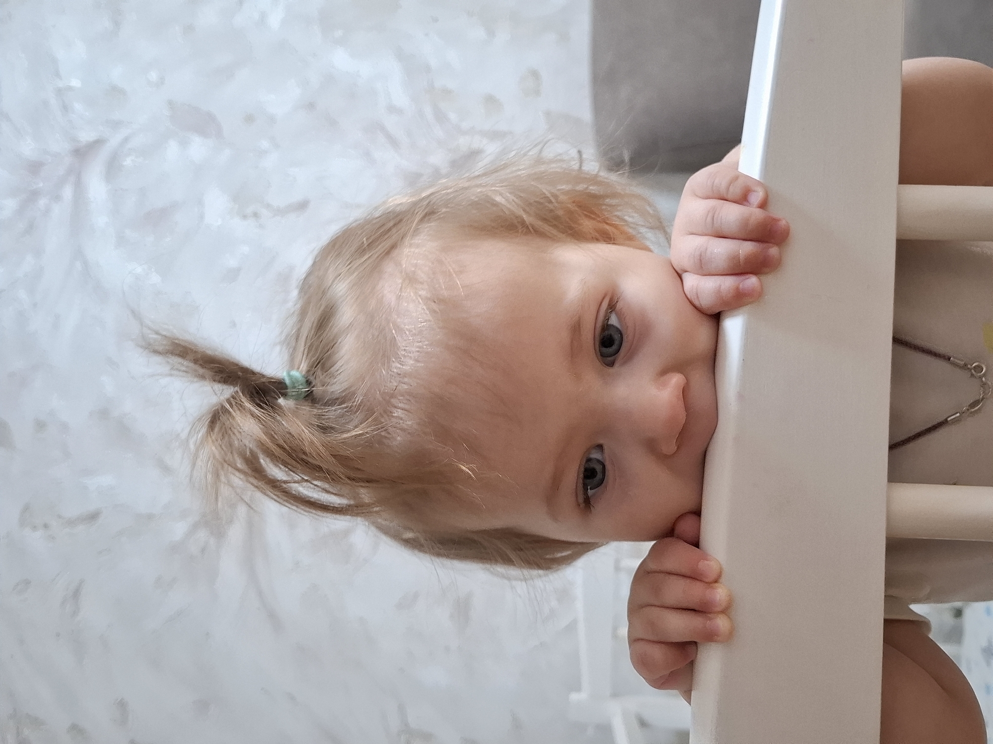 A young child with light hair tied back in a small ponytail with a green hair tie is positioned behind a white, wooden structure, possibly a high chair or crib. The child's face is visible as they peek over the structure, their skin fair and eyes appearing blue-gray. The child's hands grip the edge of the structure. The background is a textured wall with a light, possibly off-white, color. A silver chain with charms is visible on the right side of the image. This image appears to have been taken in Chaikovsky, Russia.