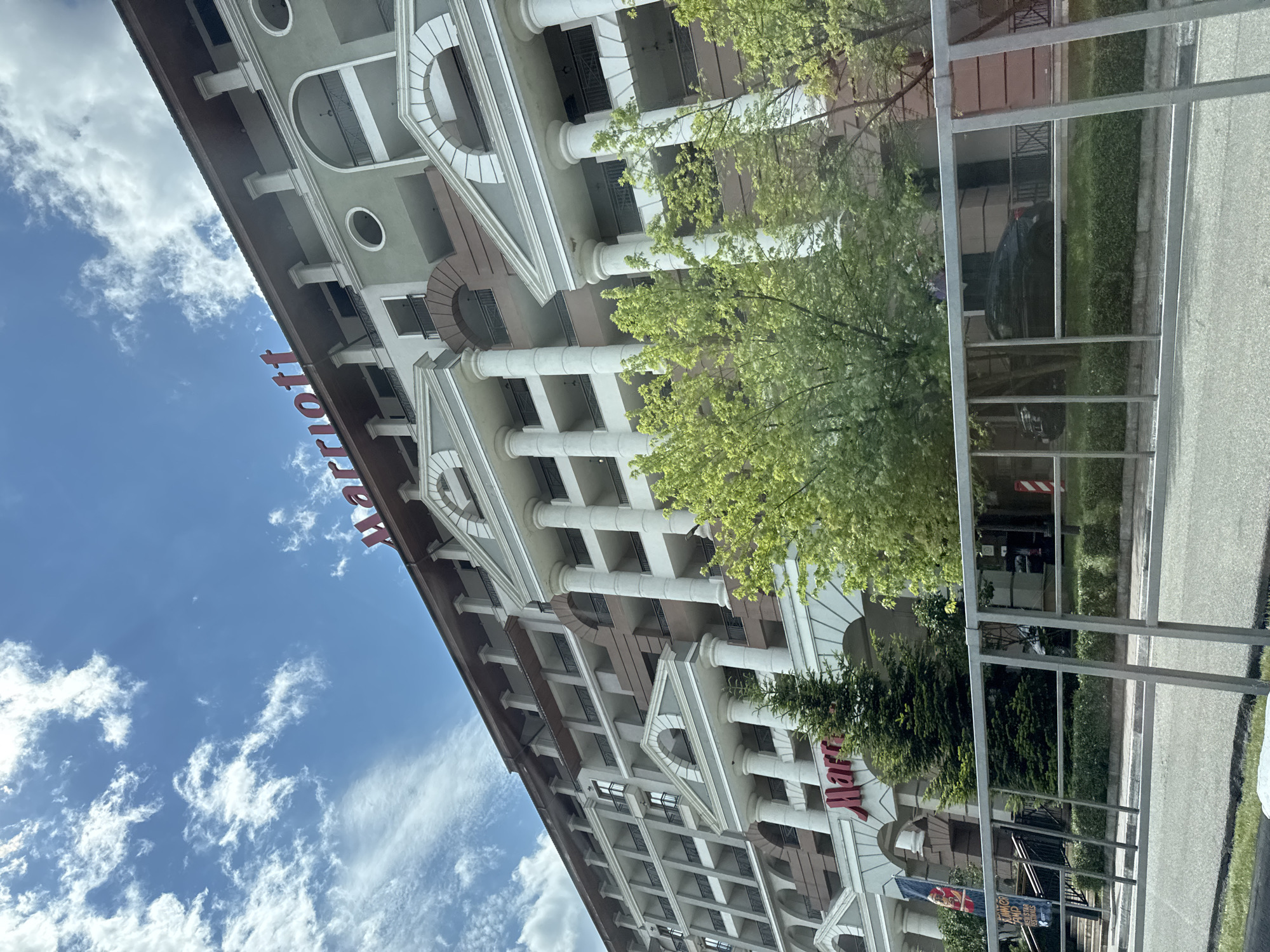 The image shows a view of the Marriott hotel in Estosadok, Russia. The hotel is a multi-story building with a light green and beige facade, adorned with white columns and balconies. The "Marriott" logo is prominently displayed in red lettering on the top of the building. In front of the hotel, there are several trees with green foliage, and a black car is visible parked in front of the building. A metal fence runs along the foreground, separating the viewer from the hotel entrance and surrounding grounds. The sky above is blue with scattered white clouds. A flag with some text and images is visible on the left side of the hotel entrance.