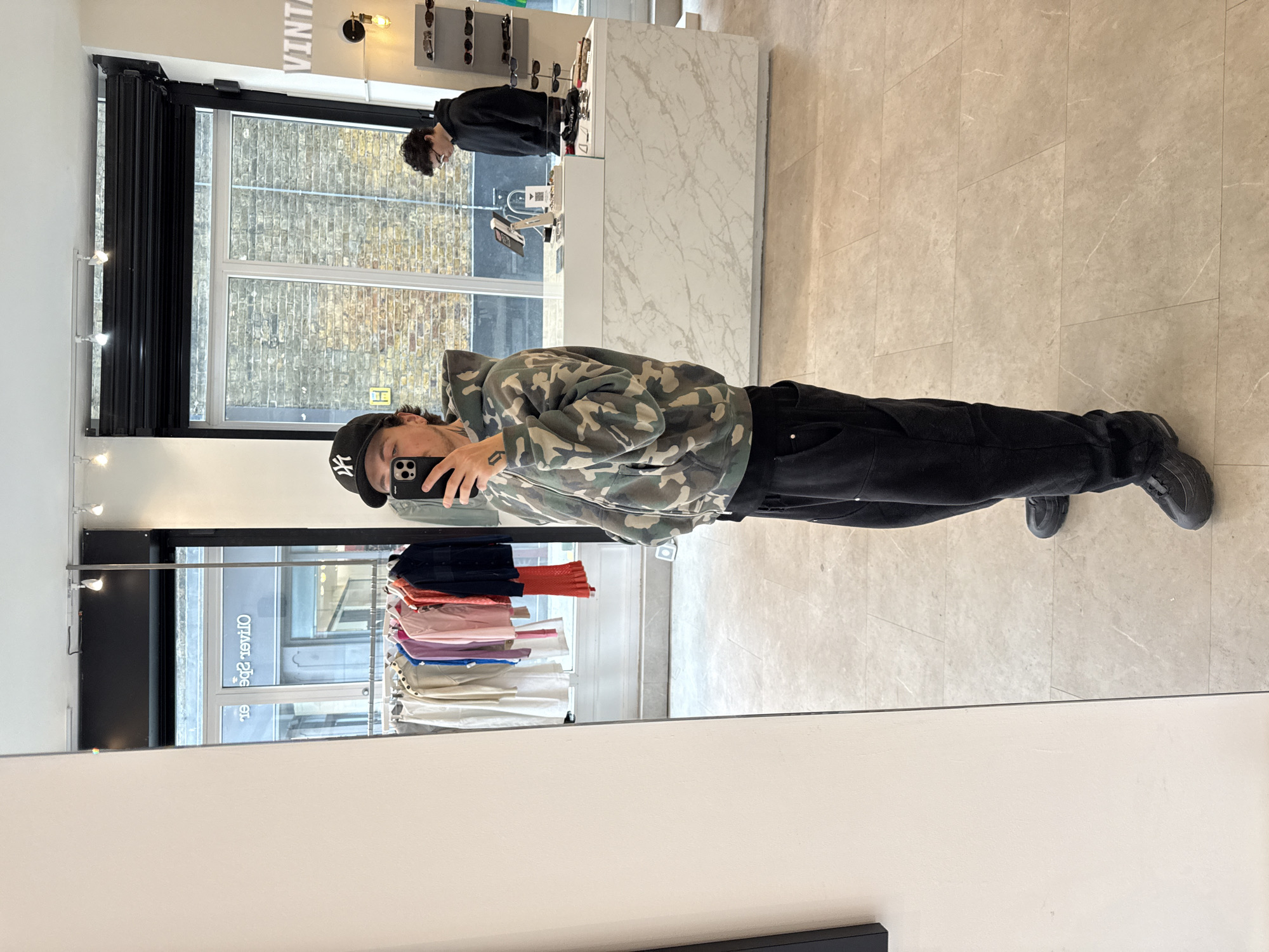 A mirrored selfie of a person in a camouflage hoodie and black pants stands in a shop in London, United Kingdom. The person is holding a black phone, wearing a black baseball cap, and facing the mirror. The shop appears to be a clothing or accessories store, with a display of clothing items visible in the mirror's reflection. Further back, a person is looking at a display case of glasses. The shop has large windows revealing a street scene with brick buildings, and the flooring is light grey tiled. The shop also features a marble-patterned counter, and vintage-style light fixtures.