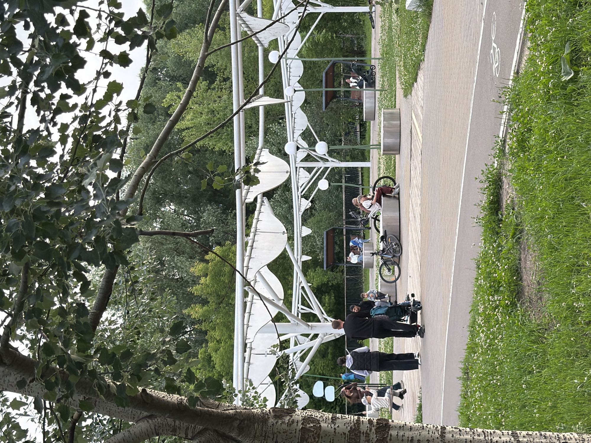 The image captures a scene in a park in Kazan, Russia, featuring a modern white canopy structure with a flowing, wave-like design. A paved path, likely a walkway or bike path, runs alongside a grassy area dotted with small white flowers. Several people are scattered throughout the scene. In the foreground, the trunk and branches of a large tree with textured bark partially obscure the view.
Underneath the canopy, two people are seated, and one person stands, possibly engaged in conversation. Further down the path, another group of people are gathered, with some sitting and others standing, some with bicycles nearby. One individual is seated on a bench, facing away from the camera, with a bicycle leaning against the bench.
A young person in a white shirt and brown pants is seated on a bench, looking down at their phone, with a bicycle parked next to them. Another person, dressed in dark clothing, is standing and facing the person on the bench. In the background, under a sheltered area, two individuals are seated on a bench, with a bicycle parked beside them.
The scene appears to be set during the daytime, with diffused lighting suggesting it could be overcast or late afternoon. The overall atmosphere seems relaxed and casual, indicative of a park setting where people are enjoying leisure time. The presence of bicycles suggests a popular spot for cycling or simply enjoying the outdoors. The modern architectural design of the canopy stands out against the natural backdrop of trees and greenery.