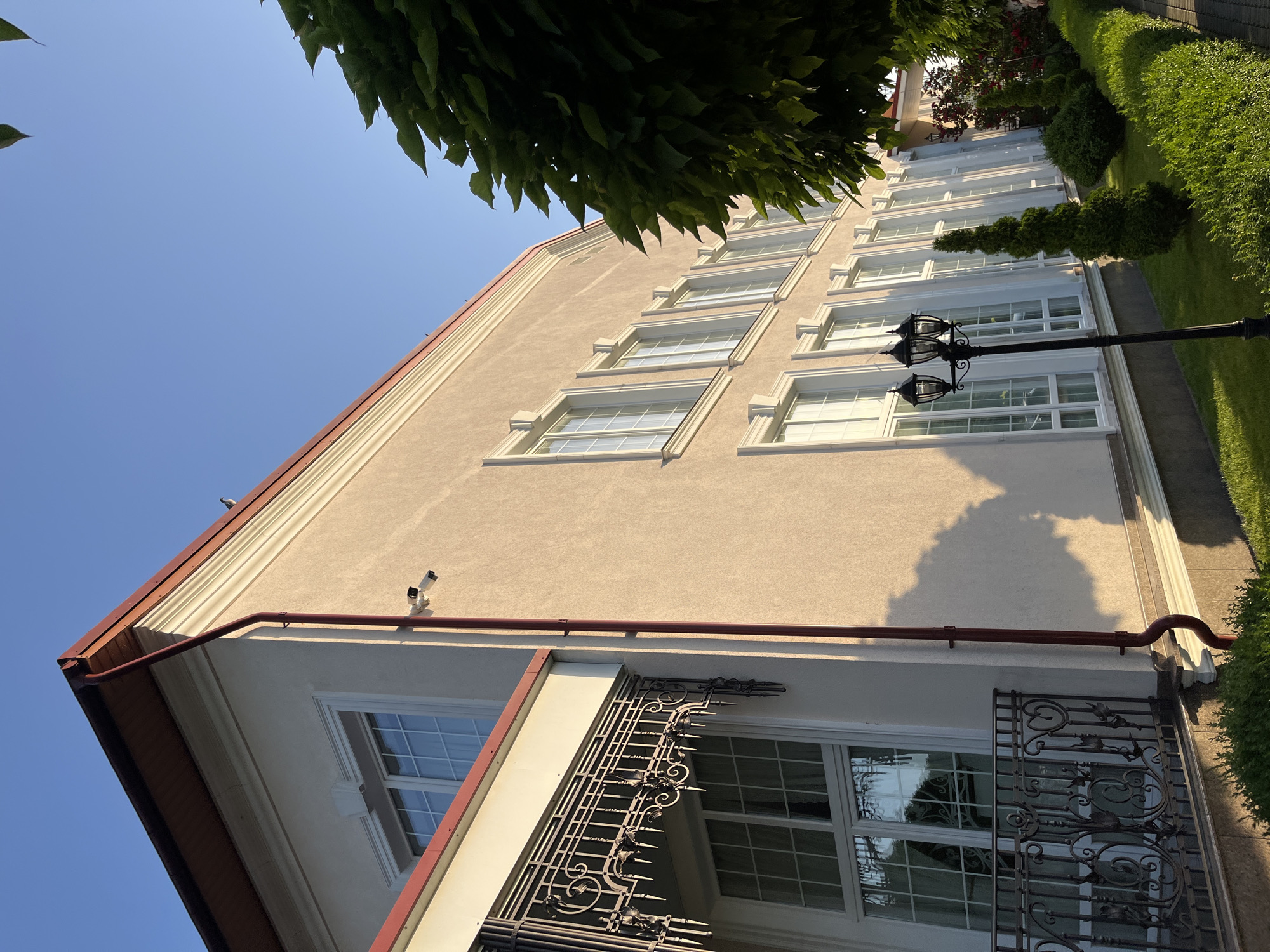 A low-angle, exterior shot captures a portion of a modern building with a light beige stucco facade, red trim along the roofline, and a series of tall, white-framed windows. A dark red downspout runs vertically along the side of the building. A security camera is mounted on the wall near the top of the downspout. To the left, an ornate wrought-iron balcony with decorative elements overlooks a large window. In the foreground, a black lamppost with three lights stands on a manicured lawn. The lawn is bordered by a neatly trimmed hedge and various sculpted evergreen bushes and a rose bush. The sky above is clear and blue. The image was taken in Dubivtsi, Ukraine, during daylight, likely in the late afternoon or early evening, as indicated by the warm lighting and long shadows cast by the building and landscaping. There are no people visible in the scene.