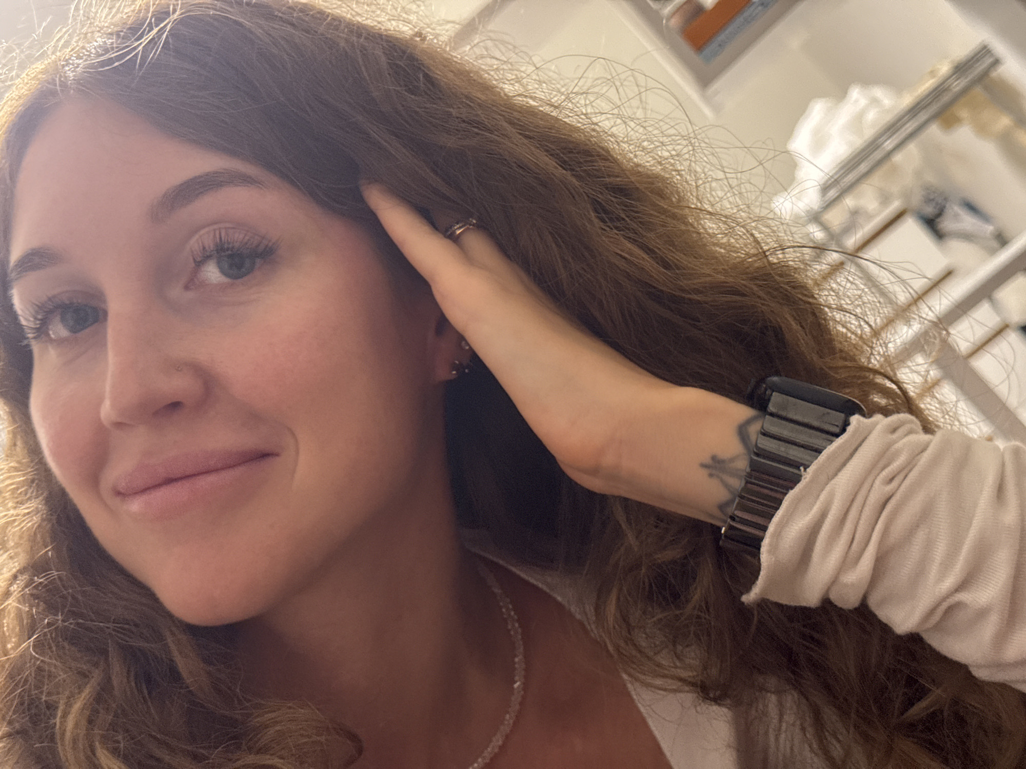 A close-up selfie shows a young woman with long, wavy brown hair, looking towards the viewer with a gentle, contented smile. Her left hand is positioned near her face, with fingers lightly touching her hair and temple. She has blue-grey eyes with prominent eyelashes, neatly shaped eyebrows, and a small stud piercing on her left nostril. Several rings adorn her fingers, and a small earring is visible in her left ear. A delicate, sparkling chain necklace is worn around her neck, resting on a light-colored, possibly off-white, long-sleeved top. On her right wrist, a dark watch with a segmented band is visible, along with a partial view of a black tattoo featuring a geometric or stylized design, possibly resembling a crown. The background is softly blurred, showing indistinct shelves or storage units, suggesting an indoor, possibly domestic, setting. The lighting appears to be artificial. The scene captures a casual, intimate moment, potentially taken in Chaikovsky, Russia. There is no visible text in the image.