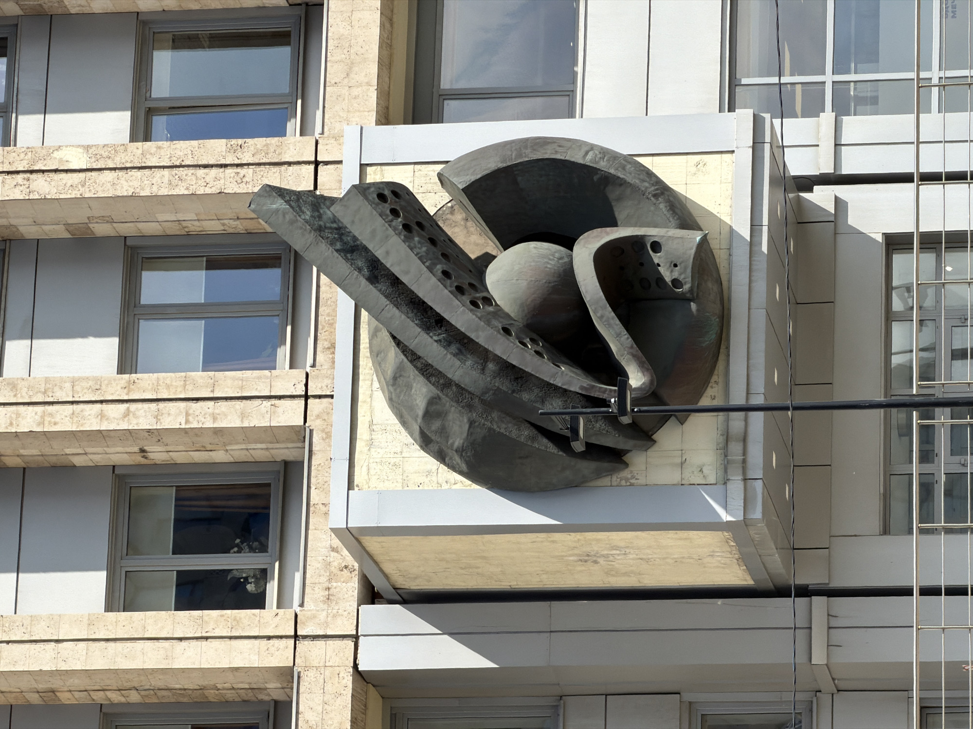 This image depicts a large, abstract metal sculpture affixed to the facade of a building. The sculpture, of dark gray or black color, features a combination of curved and angular elements, with a spherical or ovoid central form surrounded by arcing, perforated planes. The building to which the sculpture is attached is characterized by a grid of rectangular panels, some of which contain windows. The building's facade is a mix of pale stone and painted surfaces. Adjacent to the building with the sculpture is a more modern structure with a grid of windows and light-colored vertical and horizontal supports. The photograph is taken in daylight, and the city is unknown.