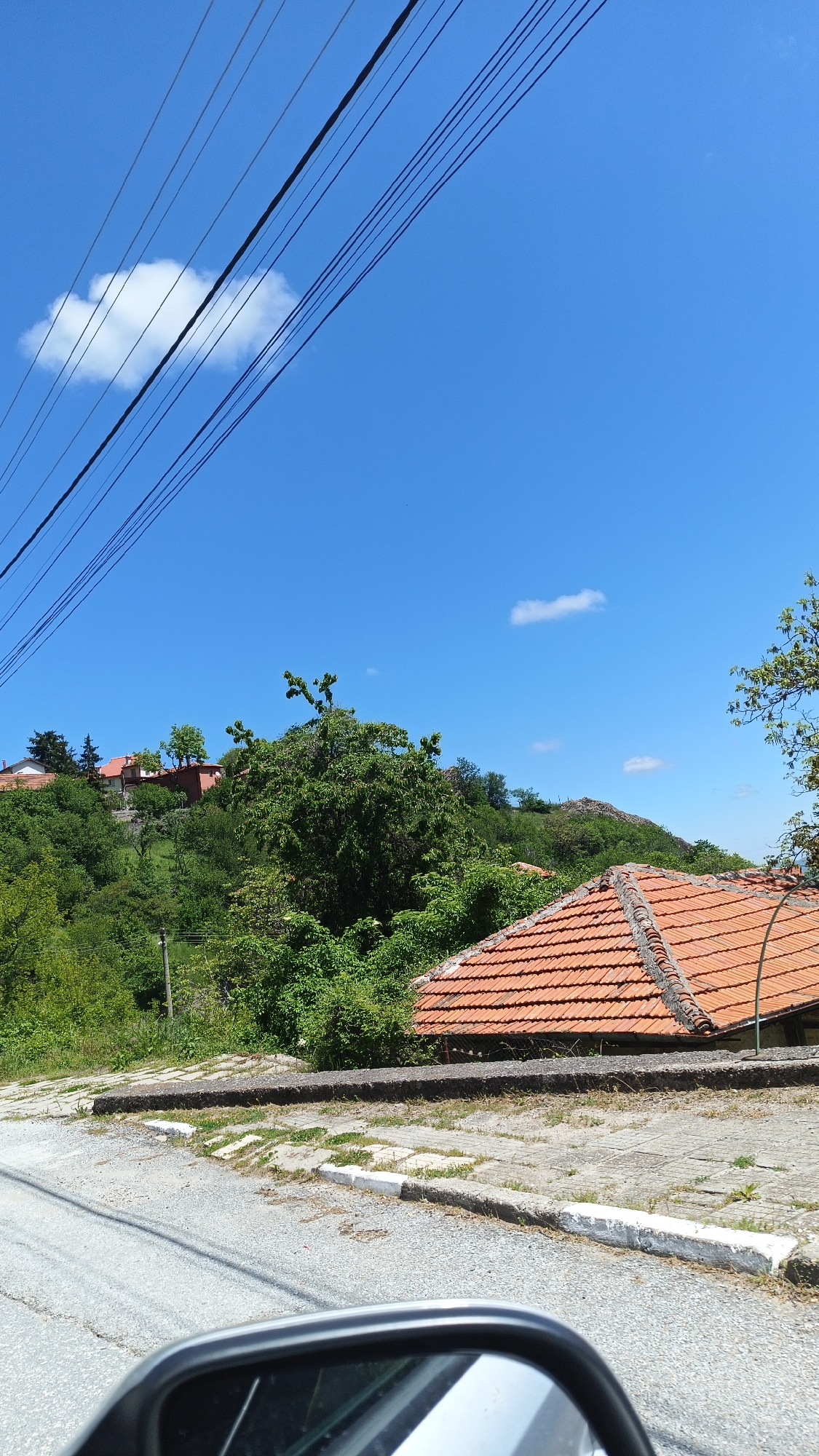 The image shows a view in Oreshets, Bulgaria, with a roadside and an upward slope. In the foreground, there is a concrete curb and paved area adjacent to an asphalt road. A side mirror of a vehicle is visible in the bottom of the frame. The midground is dominated by dense green vegetation, covering the rising terrain. The roof of a building with orange terracotta tiles is partially visible, partially obscured by the vegetation. Higher on the slope, several buildings are scattered among the trees. Above the treeline, there is a clear blue sky with wispy white clouds. Power lines stretch across the upper left portion of the sky.