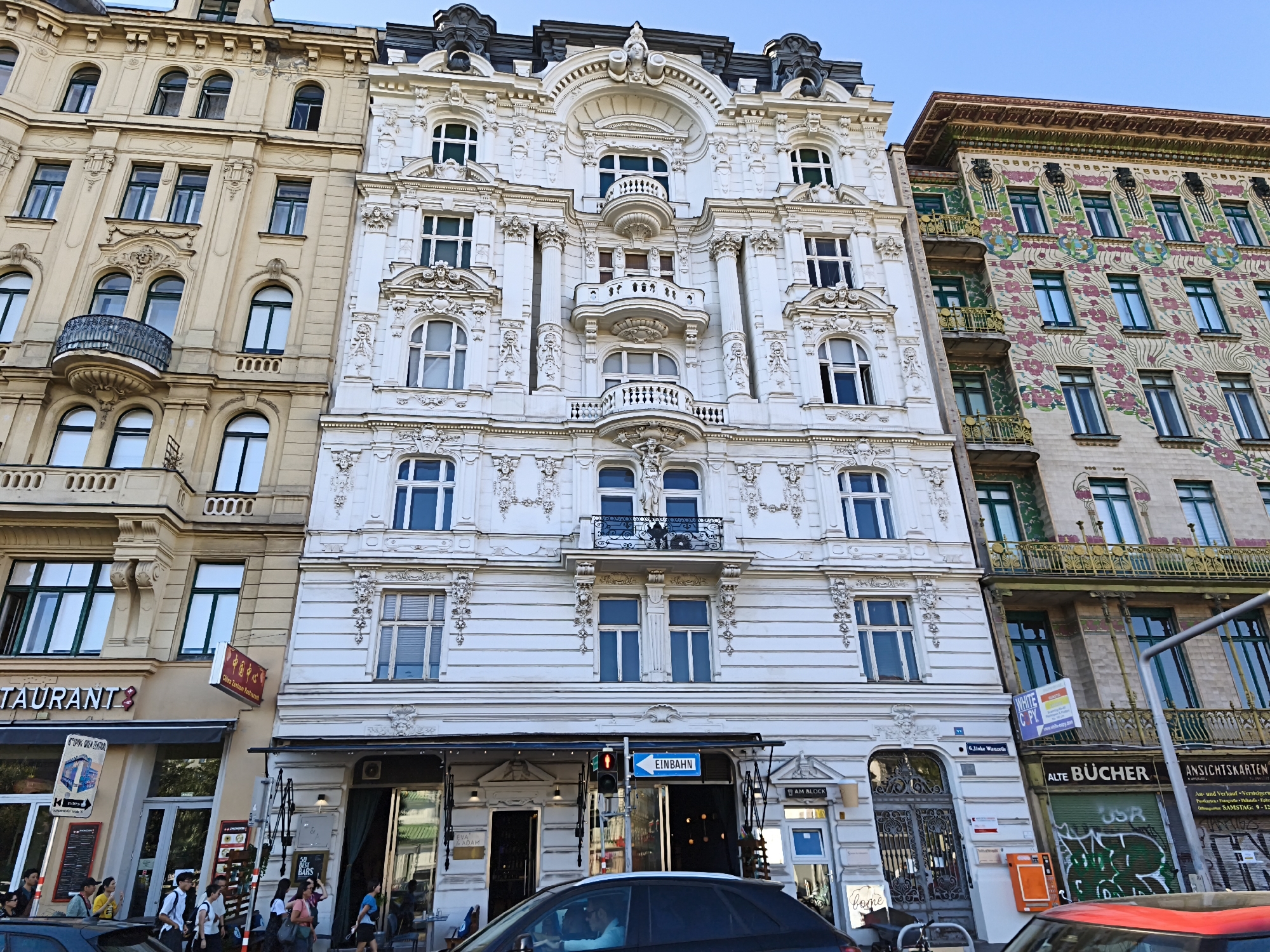 The image shows a street scene in Vienna, Austria, focusing on the facades of several adjacent buildings. The central building is a grand, white, ornate structure with numerous windows, balconies, and intricate architectural detailing, characteristic of Viennese Baroque or Art Nouveau styles. To its left is a lighter colored building with similar architectural features, and to its right is a building with a more colorful, decorative facade featuring floral motifs.
On the street level, businesses are visible. To the left, there is a restaurant with an awning and signage. To the right of the white building, there is a shop with signage indicating "ALTE BÜCHER" (old books). People are walking on the sidewalk, some entering or exiting the establishments. A traffic light is visible, with a one-way street sign ("EINBAHN") clearly displayed. A black car is parked in the foreground, obscuring part of the street. The sky above is clear and blue. The buildings are tall and imposing, with the facades displaying varying architectural and decorative styles, suggesting a historic urban environment.