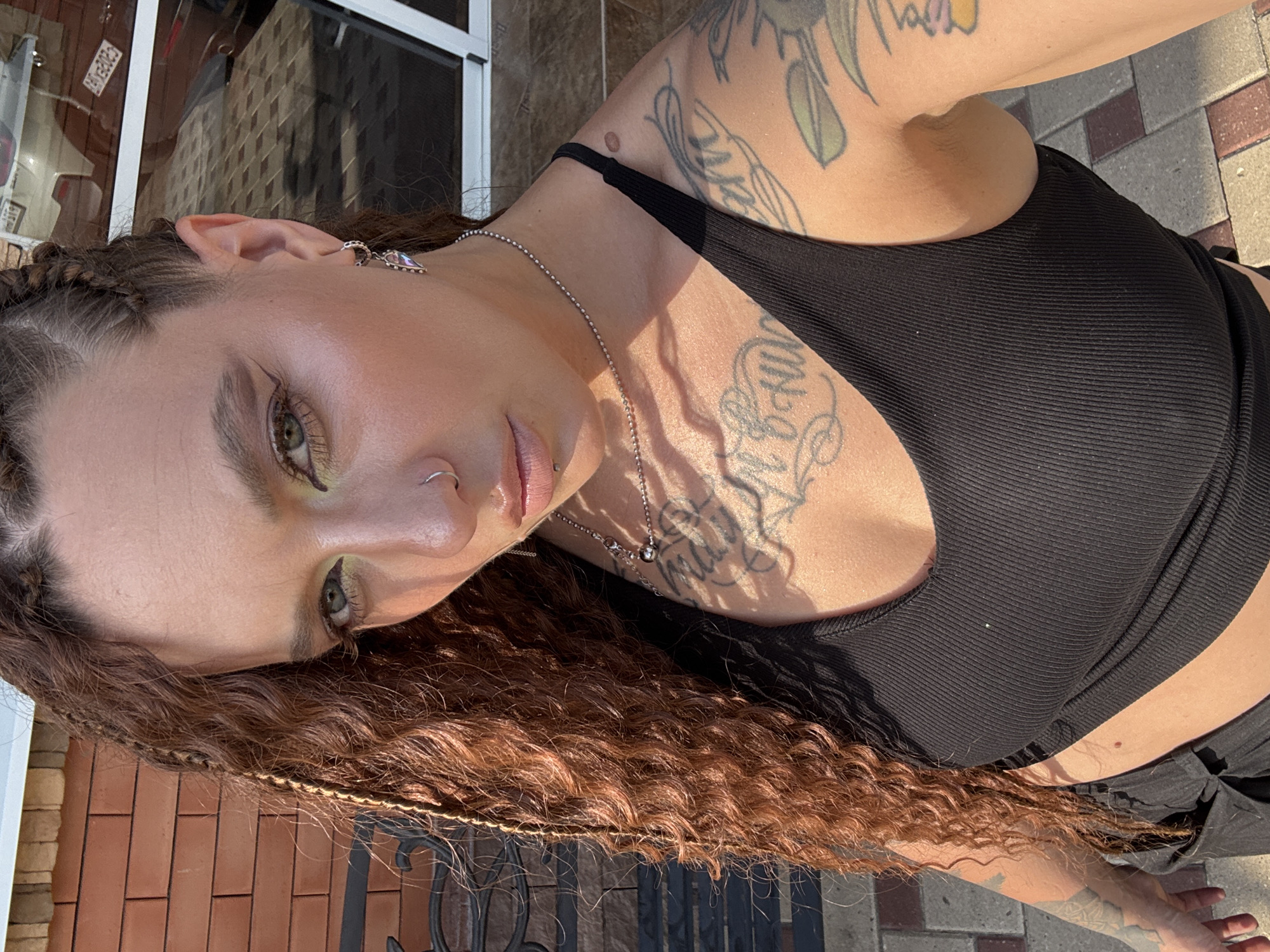 The media file depicts a person with long, curly, reddish-brown hair and distinctive braided elements near the front hairline. They are wearing a black, ribbed tank top and a silver chain necklace. The person has visible tattoos on their upper chest and arm, as well as a nose ring and an earring. Their makeup includes an application of what appears to be green eyeshadow.
The subject is positioned outdoors, with the warm sunlight casting shadows, suggesting a daytime setting, likely during sunny weather. The background includes brickwork, indicative of an outdoor patio or walkway, and a blurred building facade. The image is taken at an upward angle, focusing on the upper body and face of the person. No text is visible in the image. The overall impression is one of casual outdoor activity in a moderately urban or suburban environment.