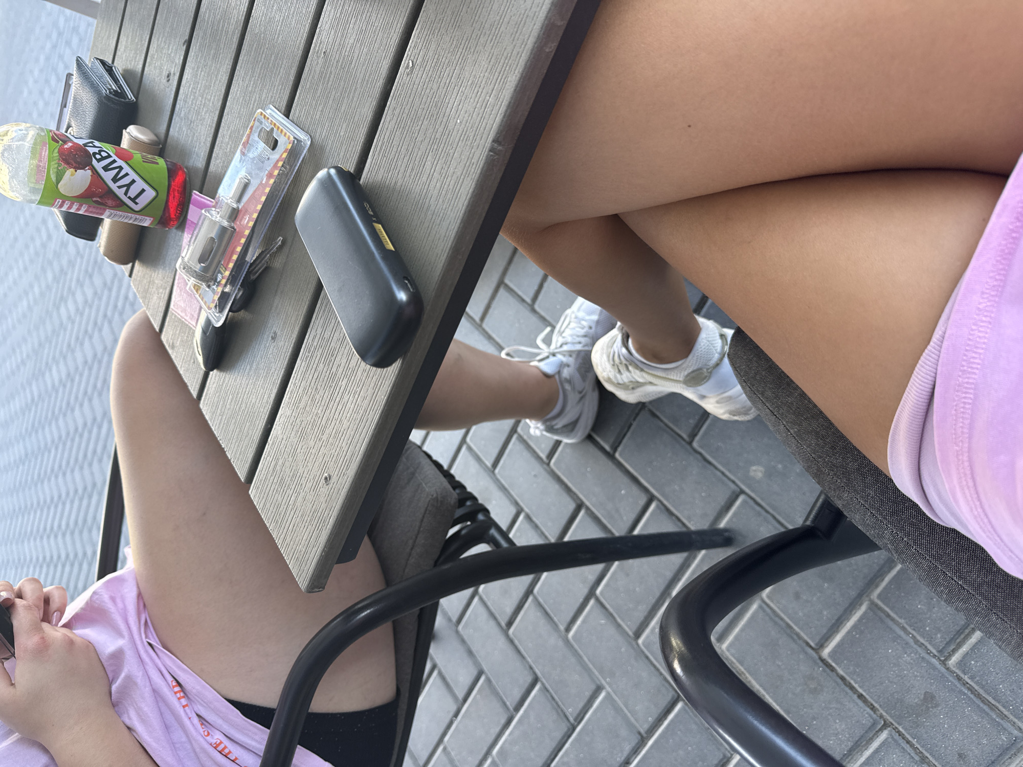 Two people are seated at an outdoor wooden table. On the table, a bottle of Tymba apple juice, a wallet, a small black electronic device, keys, and a tool are visible. One person is wearing a pink t-shirt and black shorts, and the other is wearing white sneakers and a pink skirt or shorts. The ground is paved with gray rectangular bricks. The scene appears to be in daylight, suggesting a daytime activity. There is no visible text other than on the juice bottle, which reads "Tymba" along with an image of apples. The overall setting suggests a casual outdoor gathering or meal.