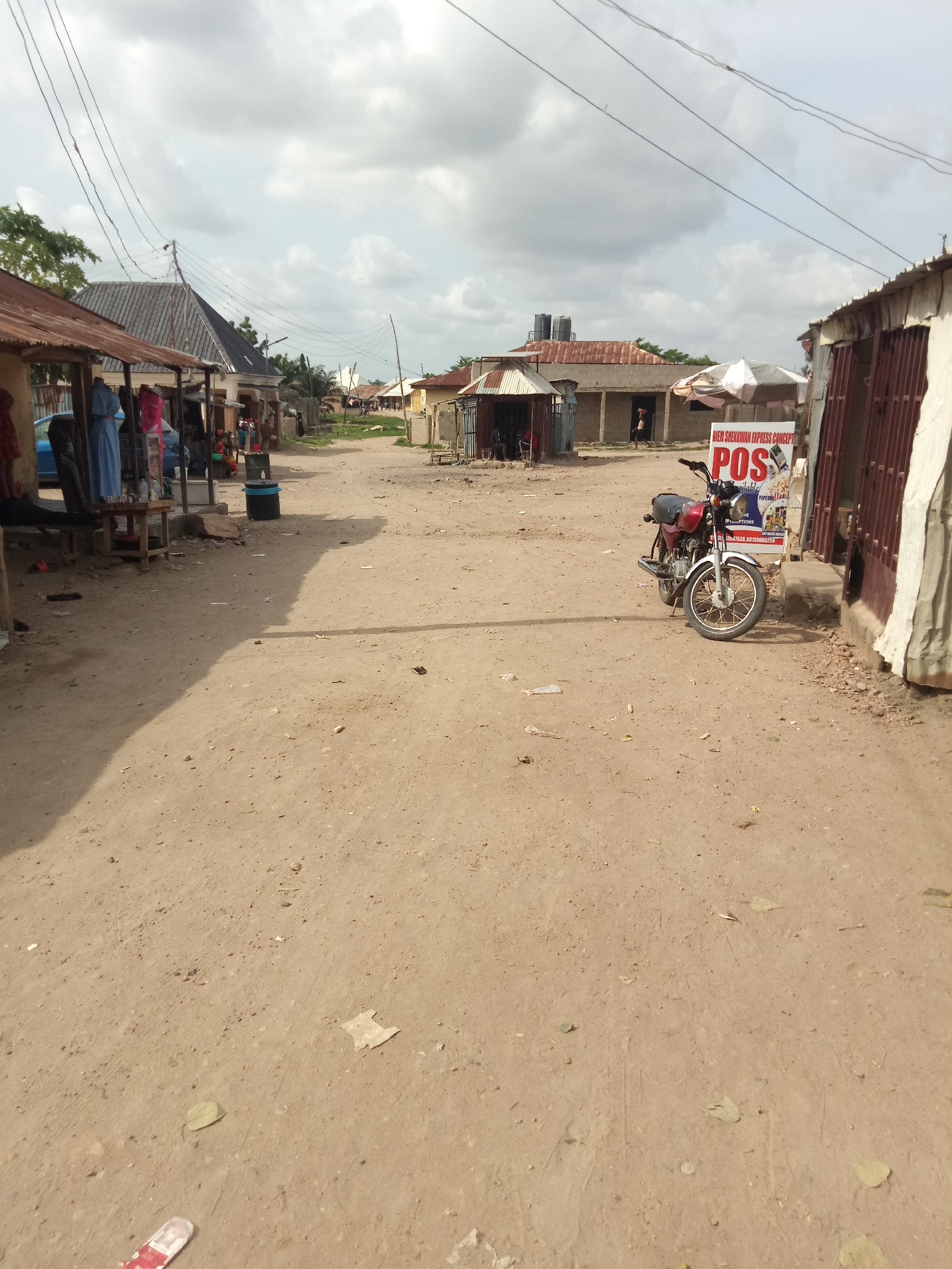 An eye-level, medium shot captures a dusty, unpaved street lined with small market stalls under a cloudy sky. In the foreground, the dirt road dominates the lower half of the frame. To the left, a stall has a corrugated metal roof and displays clothing on mannequins. Further down the street, more market stalls with similar roofing and open fronts are visible, some with people inside or near them. A blue car is parked to the left of the stalls. A dark blue trash can stands near the stalls. On the right side of the street, a motorcycle with a black seat and a red body is parked in front of a stall with a metal gate. A sign behind the motorcycle advertises "POS Available" with "BIEM SHEKKWAN EXPRESS CONCEPT" at the top. Beyond the stalls, more buildings and market structures are seen, with trees and vegetation appearing in the distance. Utility wires crisscross the sky above the scene.