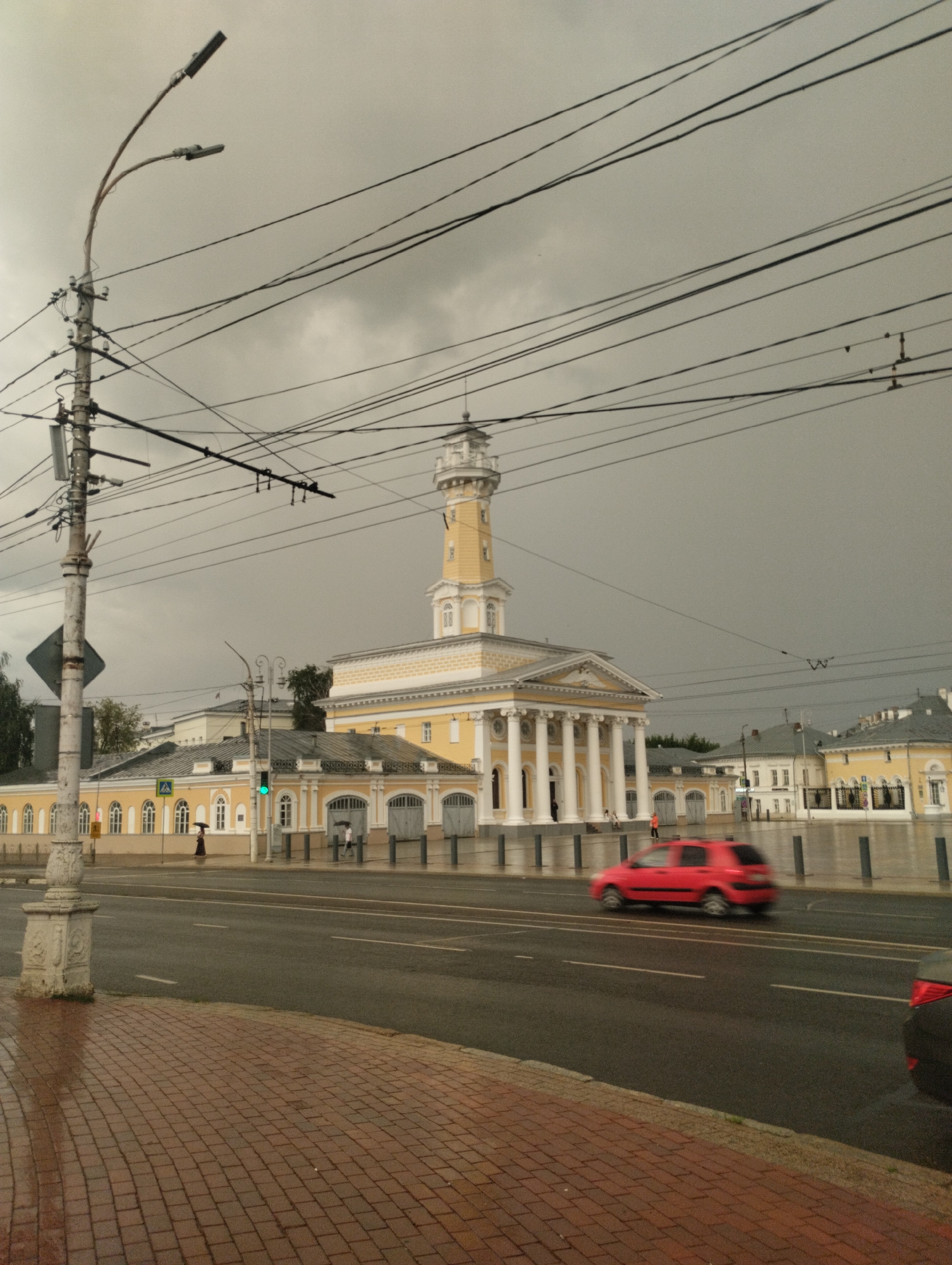 The image shows the Fire Tower in Kostroma, Russia, on a cloudy day. The tower, an iconic landmark of the city, is a prominent yellow building with white accents and a tall, slender spire. In front of the tower, a long, low building with classical columns lines the street. The street is wet, suggesting it has recently rained. A red car is captured in motion, blurred by its speed. Overhead, a complex network of electrical wires crisscrosses the sky, supported by a tall utility pole on the left side of the frame. Pedestrians with umbrellas are visible near the buildings, and a traffic light displays a green signal. The foreground features a red brick sidewalk.