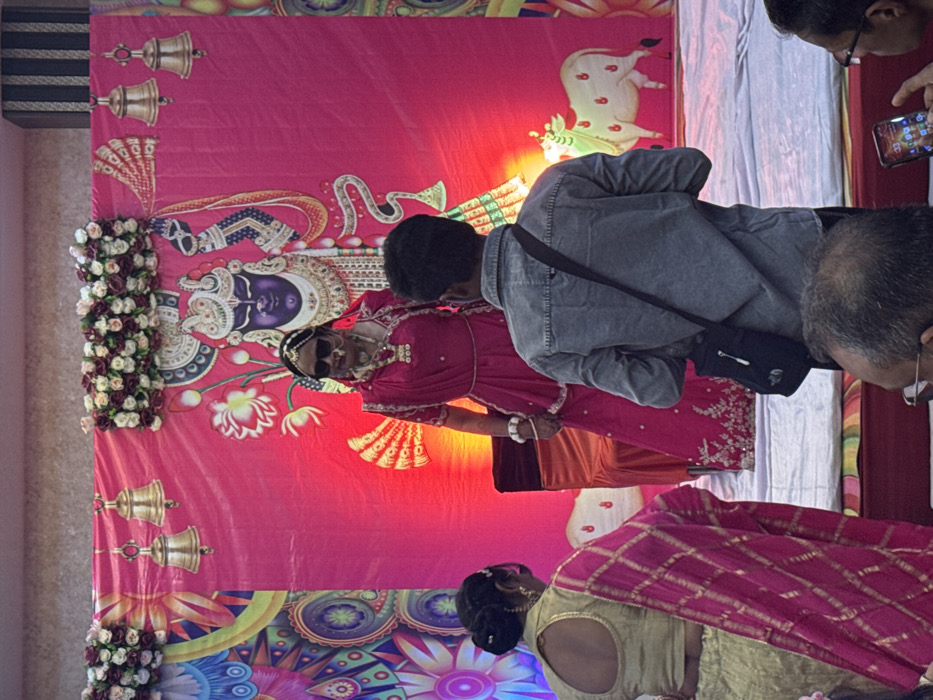 A vibrant scene unfolds with a backdrop of pink and colorful floral designs, accented by bells and a white cow illustration. A woman in a pink dress and dark sunglasses is central, seemingly part of an event or performance. A group of people, including one in a denim jacket, surround her, some capturing the moment with their phones. Another woman in a gold and pink sari is partly visible in the foreground. The event appears to be culturally significant, possibly a celebration or ceremony.