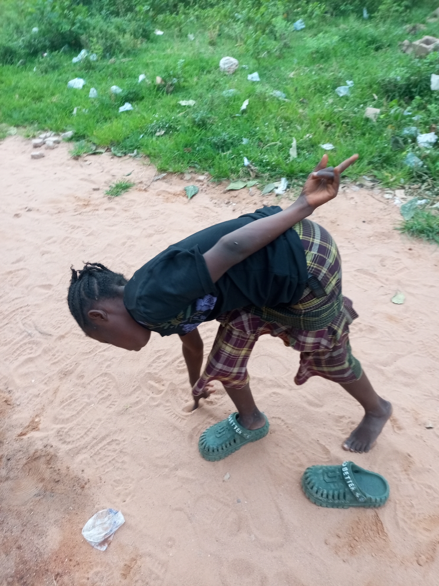 In Tempure, Nigeria, a young individual is captured in a sandy area with sparse vegetation and scattered debris. The person, wearing a black t-shirt and plaid shorts, is bent over with their right hand raised in a peace sign. One green sandal is on their left foot, while the other sandal is on the sand nearby. The ground is composed of fine sand, with faint footprints visible. A crumpled plastic bag lies on the sand in the foreground. The background consists of overgrown green grass, with several small pieces of white plastic waste visible.