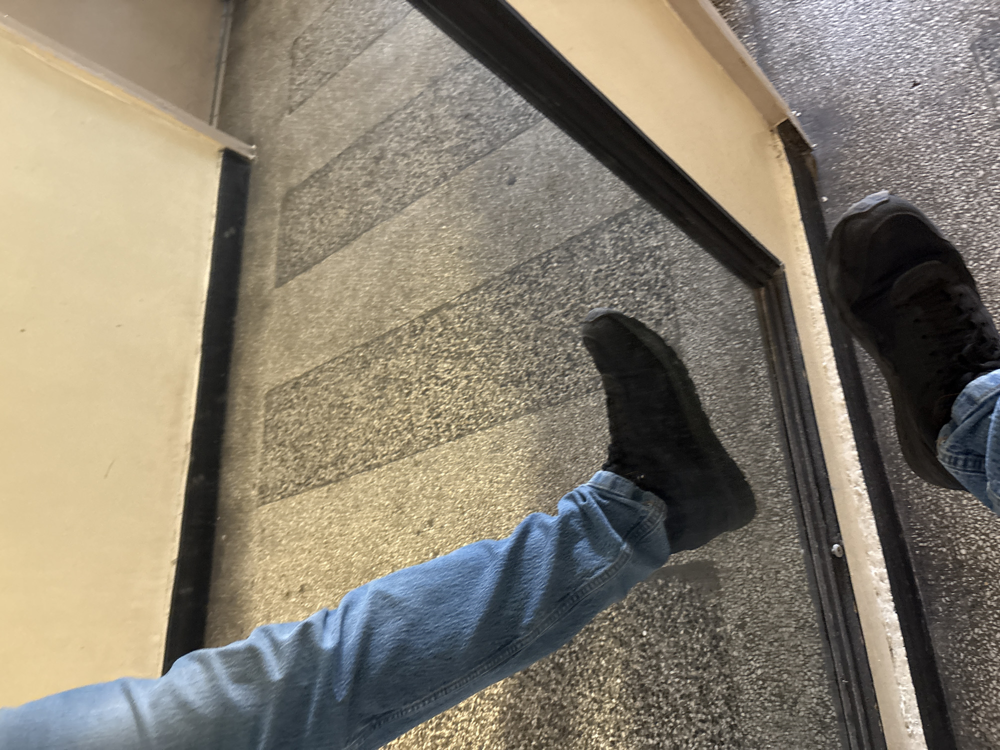 The image shows a person's legs and feet wearing blue jeans and black sneakers. The person is standing on a speckled floor, possibly a hallway or an interior common area. There is a reflective surface, likely a mirror or polished metal, in the foreground, showing the person's reflection. The wall above the floor appears to be painted a light cream color.
The image was taken in Plovdiv, Bulgaria. The specific location is the entrance area of the Plovdiv Regional Ethnographic Museum, located in the Old Town of Plovdiv. The mosaic flooring and the architectural style of the wall are characteristic of this historic building.