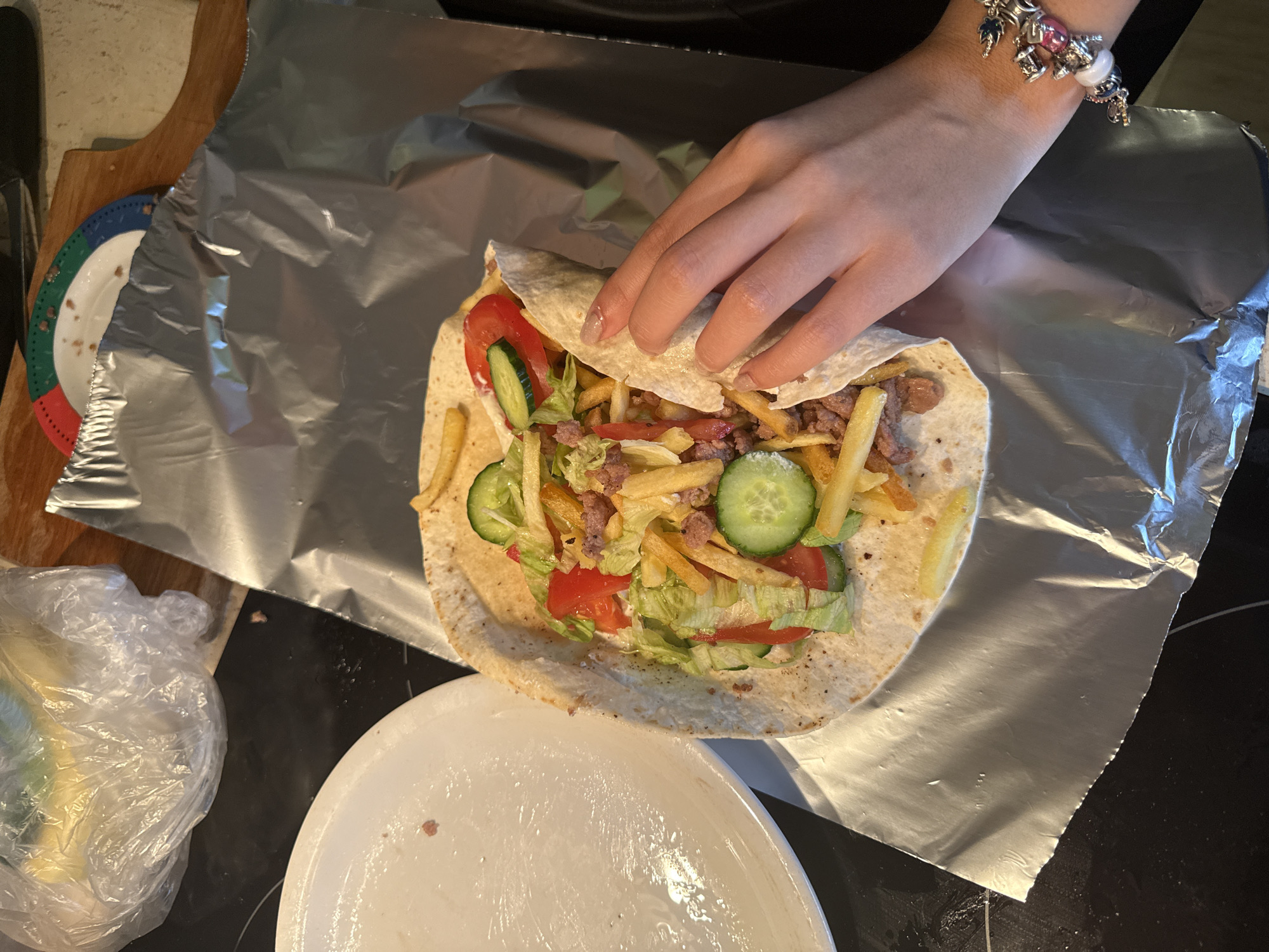 A hand with a bracelet is shown wrapping a burrito. The burrito is filled with lettuce, tomatoes, cucumbers, french fries, and meat. The burrito is placed on a piece of aluminum foil on a black surface. A white plate is visible to the left of the burrito. In the background, there is a wooden surface and a plastic bag containing yellow items. The scene is a kitchen counter, likely in a residential setting in Stara Zagora, Bulgaria, based on the provided context.
