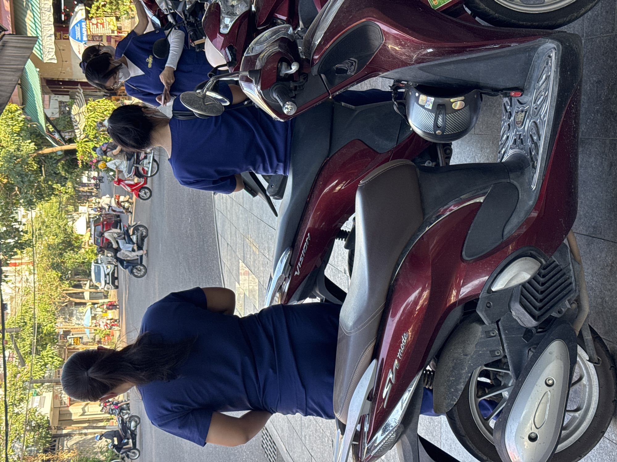 The image captures a street scene in South Quy Nhon Ward, Vietnam, likely during daytime with ample sunlight creating shadows. In the foreground, two individuals wearing dark blue uniforms are partially visible, with their backs to the camera. They appear to be standing on a sidewalk next to several parked motorcycles. The motorcycles are dark red and appear to be scooters, with one having a "VISION" logo visible on its side panel. Another motorcycle has an "SH mode" logo. A black helmet with a carbon fiber pattern is hanging from one of the motorcycles.
In the background, the street is moderately busy with vehicular traffic, including motorcycles and cars. Several people are seen on motorcycles, some wearing helmets and masks. Buildings line the street, with awnings and greenery suggesting a commercial or residential area. The overall impression is one of everyday urban life in a Vietnamese city.