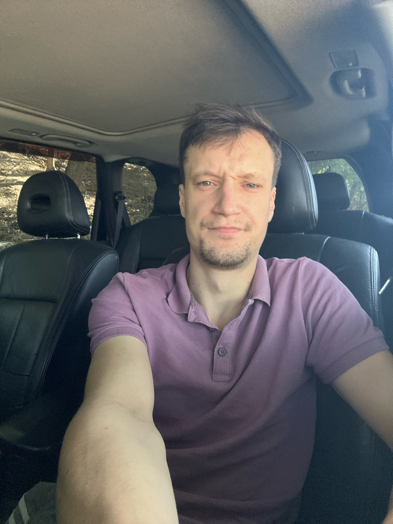 A man with short brown hair and a goatee is sitting in the driver's seat of a car, looking at the camera with a slightly furrowed brow and a neutral expression. He is wearing a purple polo shirt. The car's interior is visible, including black leather seats and a sunroof. The background appears to be a natural setting with trees. The image was taken in Makiv, Ukraine.