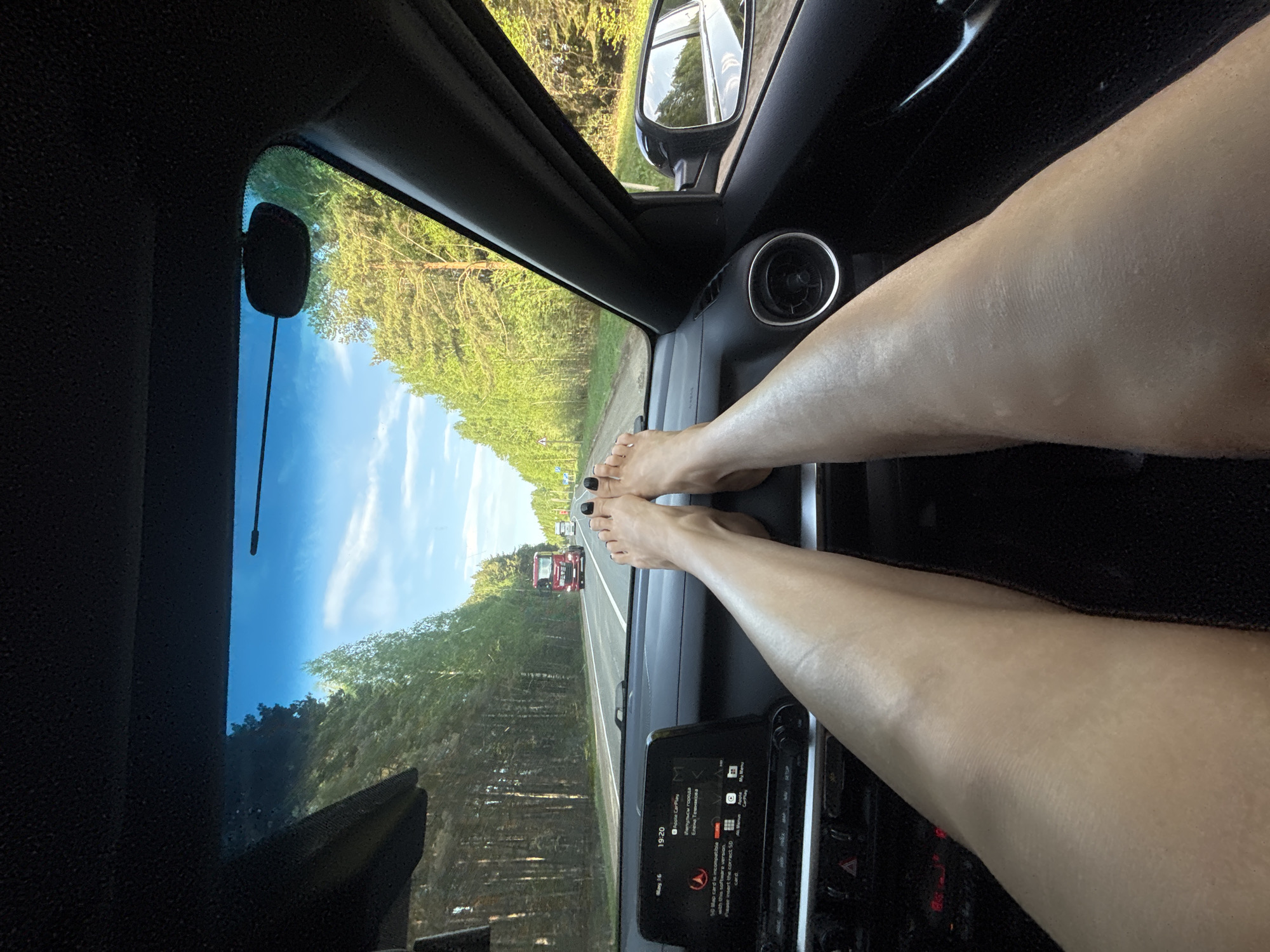 The image shows an interior view from the driver's seat of a car traveling down a road flanked by trees, presumably in a rural area. A portion of the windshield and side mirror are visible, revealing a blue sky and greenery. A truck is visible on the road ahead.
The distinguishing feature of the image is a pair of legs and feet placed on the dashboard of the vehicle, obscuring the car's infotainment system and parts of the interior. The toenails are painted black.
There is text indicating that the image was taken on May 16th at 19:20. It indicates that a SD card is incompatible. The image does not contain any details that are specific to Uspenskii, Russia.