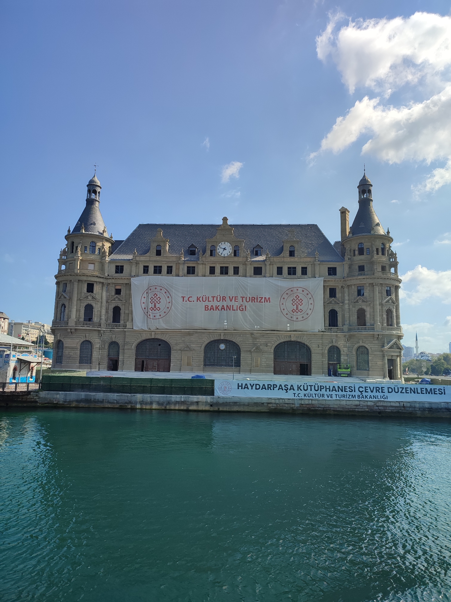 The image captures a large, ornate building with distinctive architecture, possibly a historical landmark, situated on the waterfront in Kadıköy, Turkey. The building features multiple towers with conical roofs, arched windows, and decorative stonework. A large white banner is prominently displayed across the facade, bearing text in Turkish and a central emblem. Below the building, a lower banner with similar text is also visible. The water in the foreground is a deep teal color, with ripples indicating movement. The sky above is a clear blue, punctuated by scattered white clouds. In the background, other buildings and structures can be seen, as well as distant trees and a minaret. The scene appears to be a daytime, outdoor shot, likely taken from a boat or the opposite side of the water.