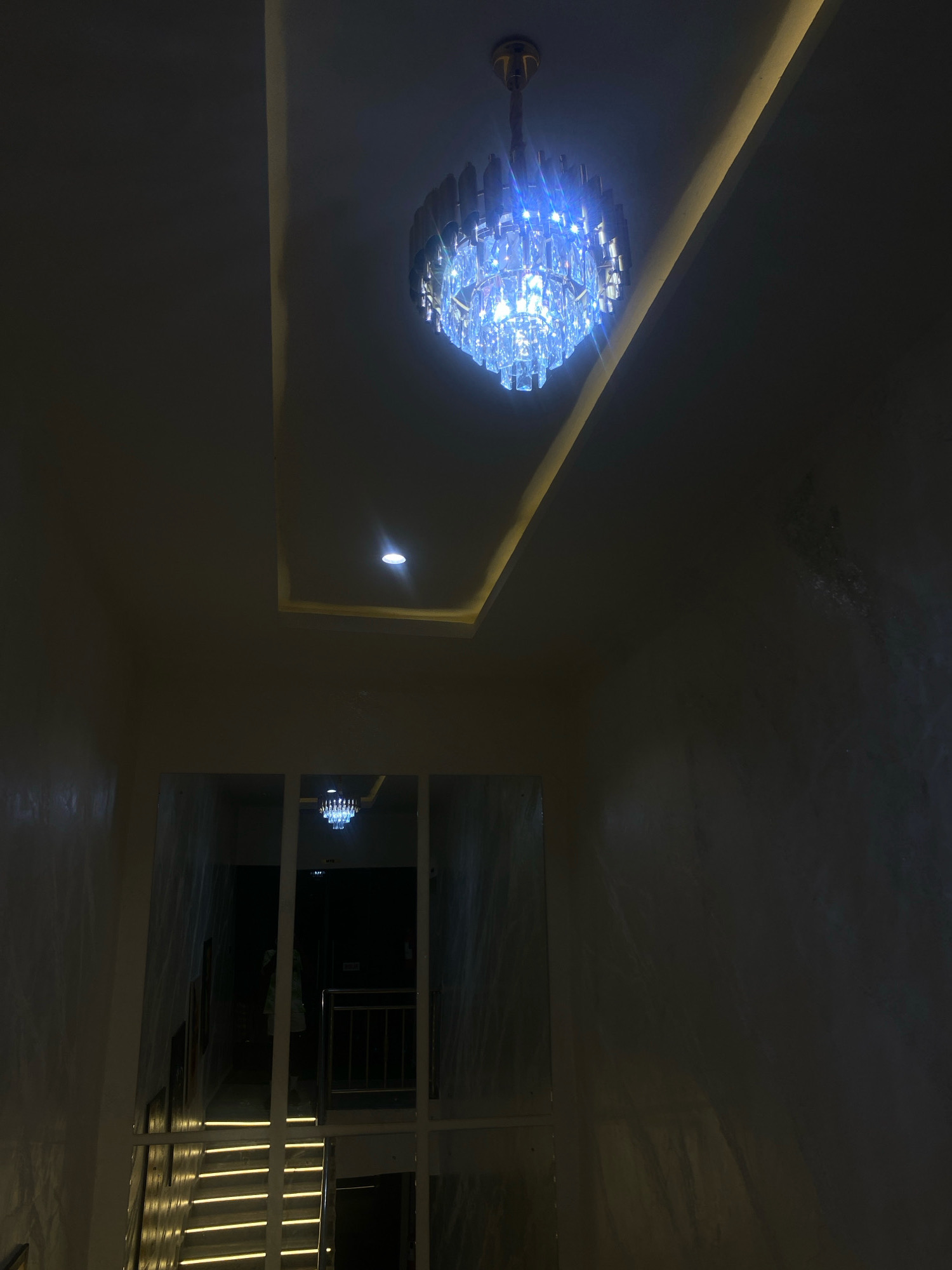 The image depicts a modern interior space, likely a hotel or residential building, with a focus on architectural lighting. A prominent crystal chandelier with blue-toned illumination hangs from the ceiling, which features recessed lighting along an angled border. The overall ambiance is dim, with the chandelier and other light sources providing the primary illumination.
In the foreground, a staircase with illuminated risers is visible, suggesting a vertical transition within the building. The walls appear to have a textured finish, contributing to the contemporary aesthetic. Further into the background, a hallway is visible, with another similar chandelier reflected in what appears to be a mirrored surface or a glass partition. The scene is devoid of any discernible human activity or specific identifying landmarks that would pinpoint its location within Barden Yana, Nigeria. The image is primarily a study of light and architectural design elements.
