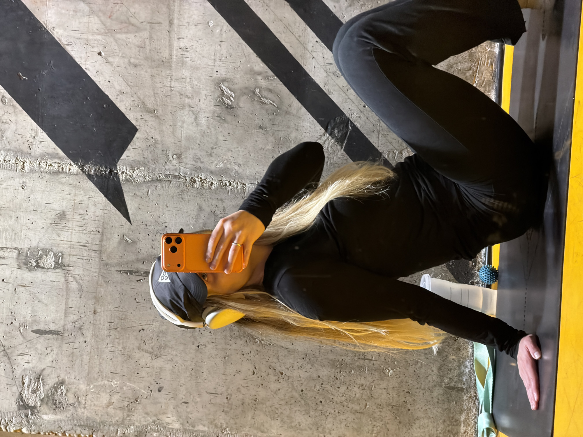 The image, rotated 90 degrees to the left, depicts a person lying on a concrete floor in what appears to be an indoor fitness or workout space in Tel-Aviv, Israel. The subject has long blonde hair and is dressed in a black long-sleeved top and black pants. They are wearing a black cap and white over-ear headphones. The person is holding an orange smartphone, possibly an iPhone, with their left hand, which has a ring on a finger, suggesting they are taking a selfie.
The concrete floor features bold black diagonal stripes, and some sections show a rough, textured finish. The person is situated next to a bright yellow mat or border. Near their outstretched arm on the yellow mat, a clear plastic cup with a lid, resembling a shaker bottle, is visible alongside a small blue spiky ball, likely a massage ball. A light green strap or band also lies on the yellow surface. No visible text is present in the image.