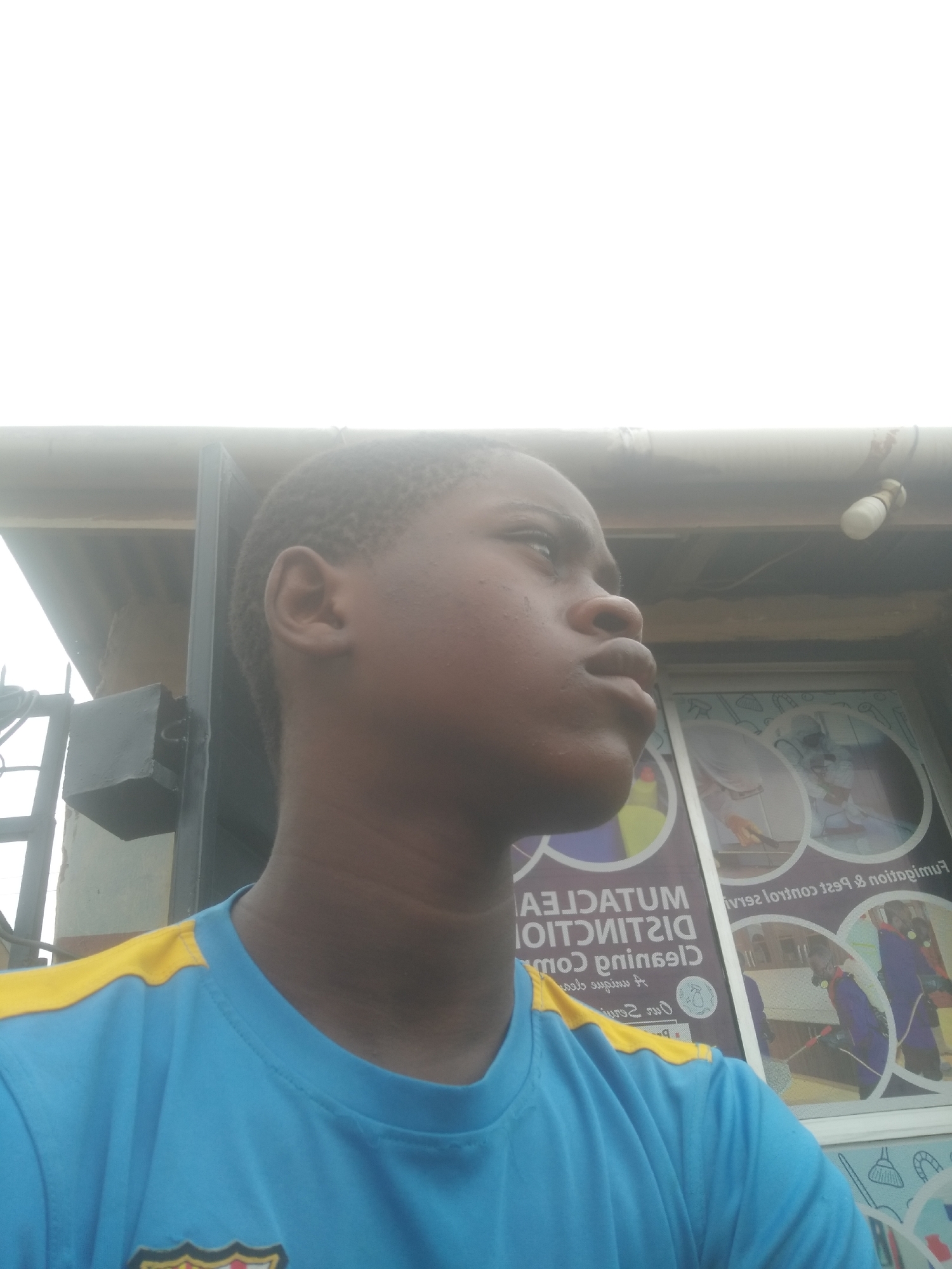A young person with dark skin and short hair is looking upwards. They are wearing a light blue t-shirt with yellow accents. Behind them are signs for a cleaning and pest control company, indicating the location is likely in an urban area. A building and overhead wires are also visible in the background. The location is assumed to be Shangisha, Nigeria, based on the prompt.