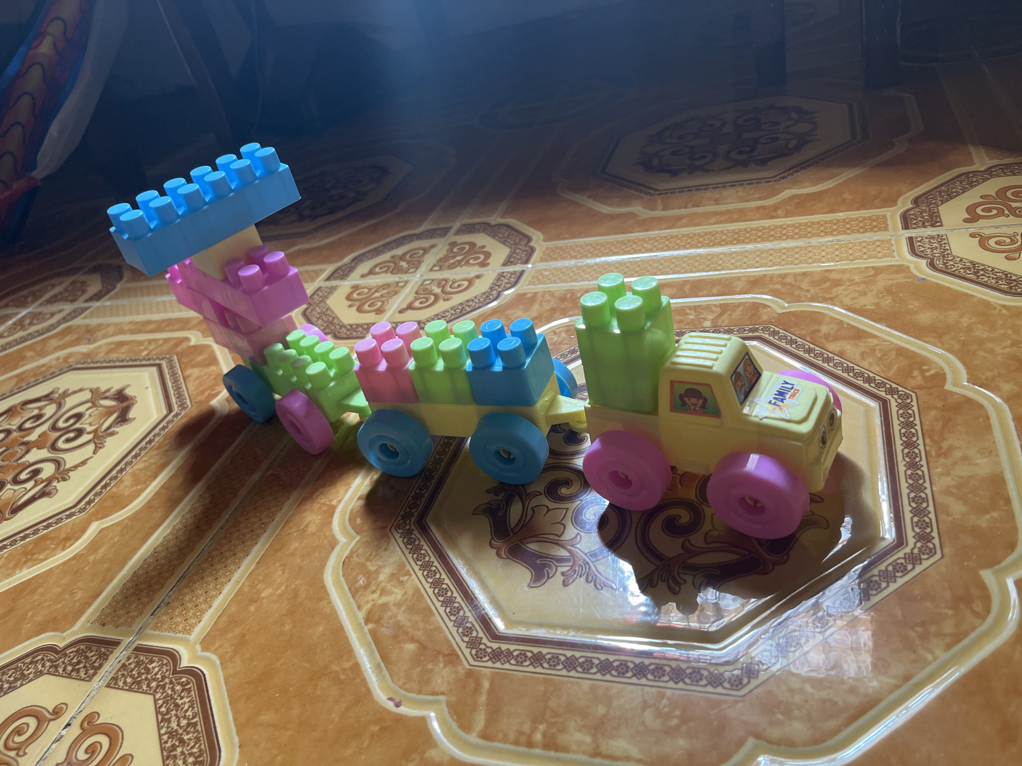 In Bumi Mulyo, Indonesia, a toy train made of colorful building blocks sits on a patterned tiled floor. The train consists of a blue, pink, and green engine, followed by several colorful train cars. The engine has a yellow cab with a sticker that says "FAMILY". The train is positioned diagonally across the frame, with the engine on the right and the last car on the left. The floor tiles are orange and cream colored with decorative borders. Sunlight casts shadows on the floor, indicating an indoor setting with artificial lighting. There are no people visible in the image.