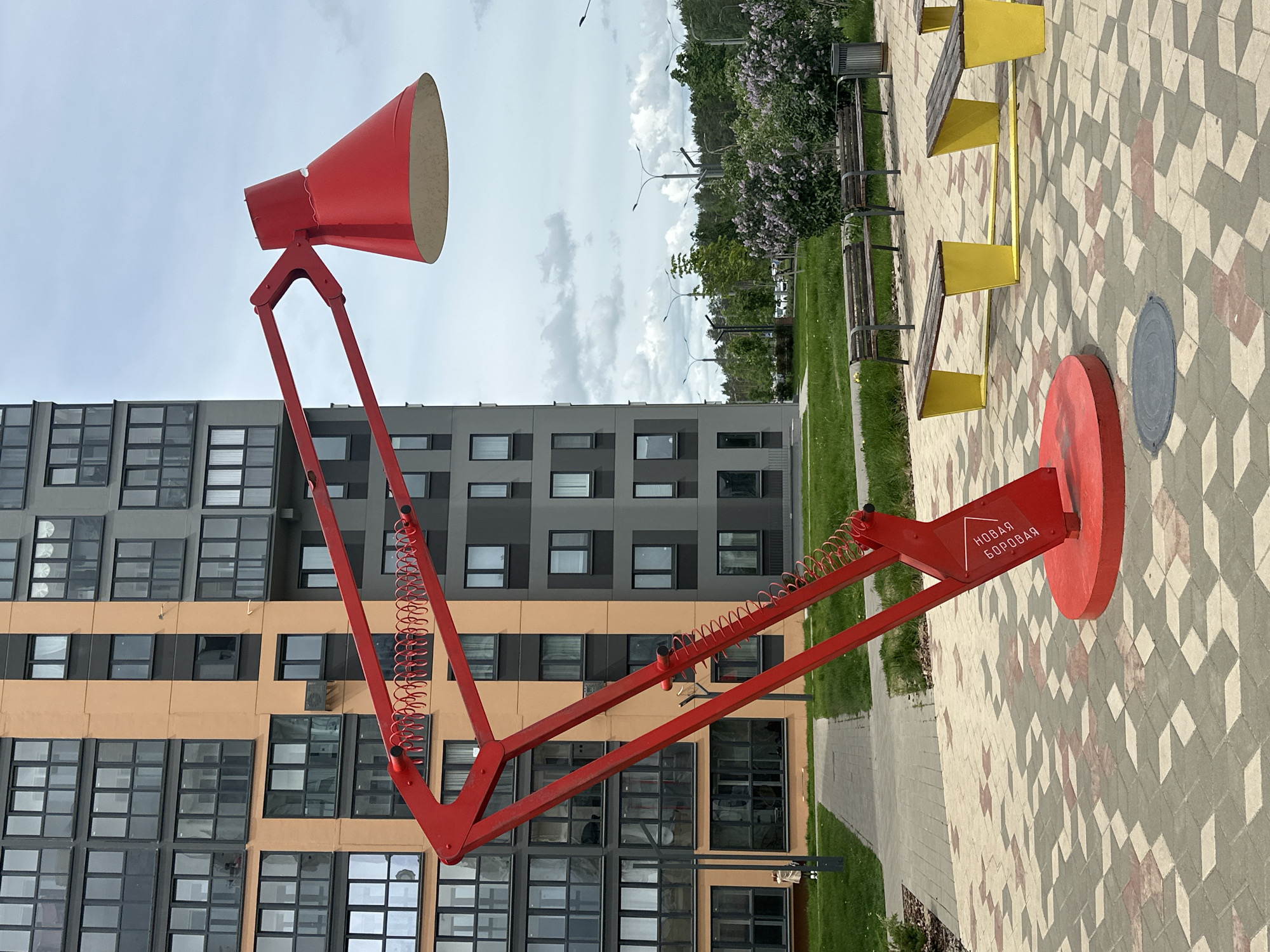 The image shows a large, red, articulated lamp sculpture in Бараўлянскі сельскі Савет, Belarus. The lamp stands on a red circular base and features a red conical lampshade. The sculpture is positioned on a paved area with a geometric pattern. Behind the lamp is a modern building with multiple windows and a color scheme of gray and orange. A patch of grass is visible to the right of the lamp, along with park benches that are yellow and wood. Trees and flowering lilac bushes are in the background, with a cloudy sky above.