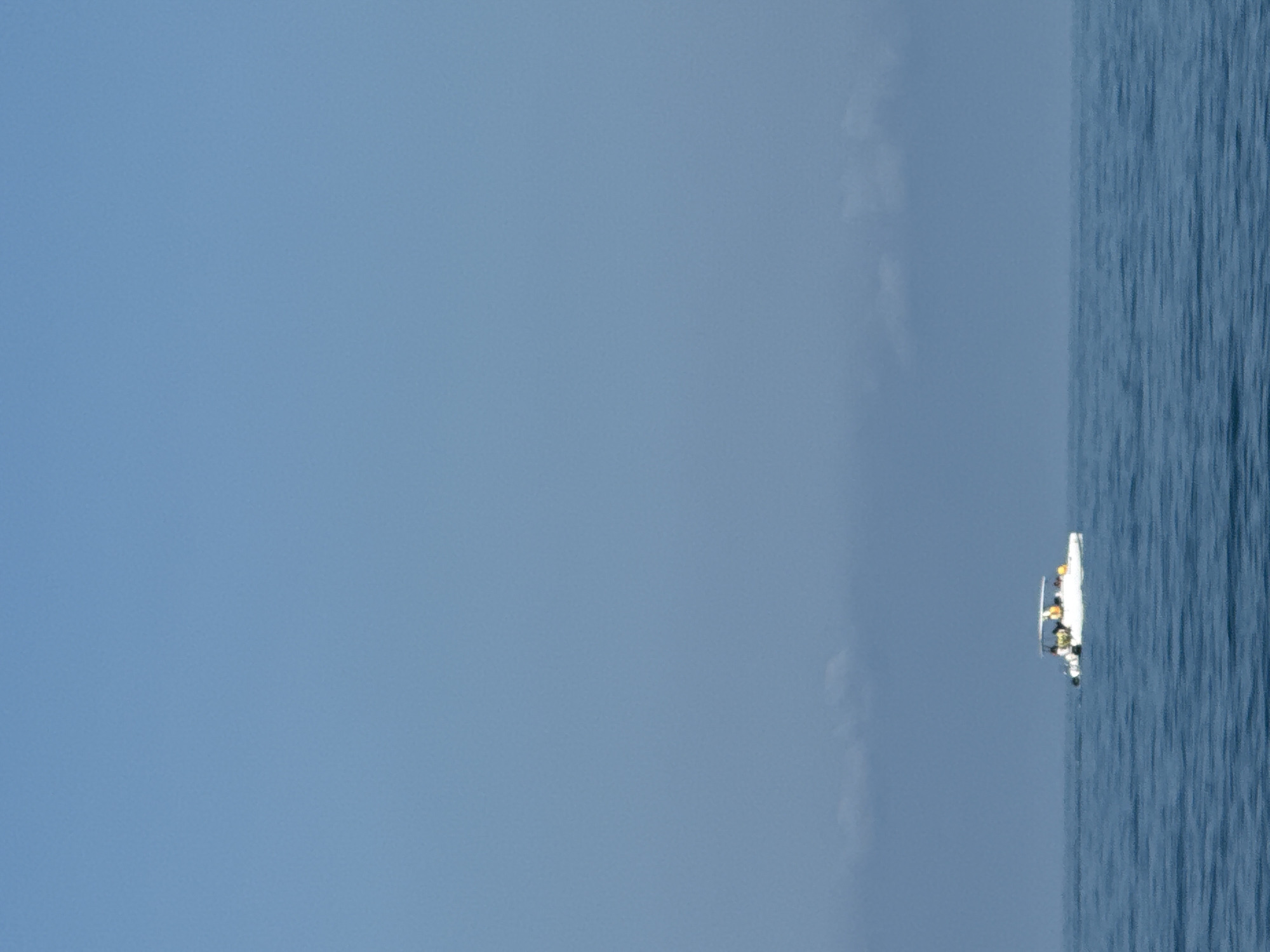 A small white boat with a canopy is visible on a vast expanse of calm blue water under a clear, pale blue sky. The boat appears to have several people on board, some of whom are wearing yellow life vests. The horizon line is indistinct due to atmospheric haze, with faint clouds near the water. The scene suggests an open ocean or large body of water. The image does not contain any elements that would place it specifically within the city of Sochi, Russia, or any other urban environment.