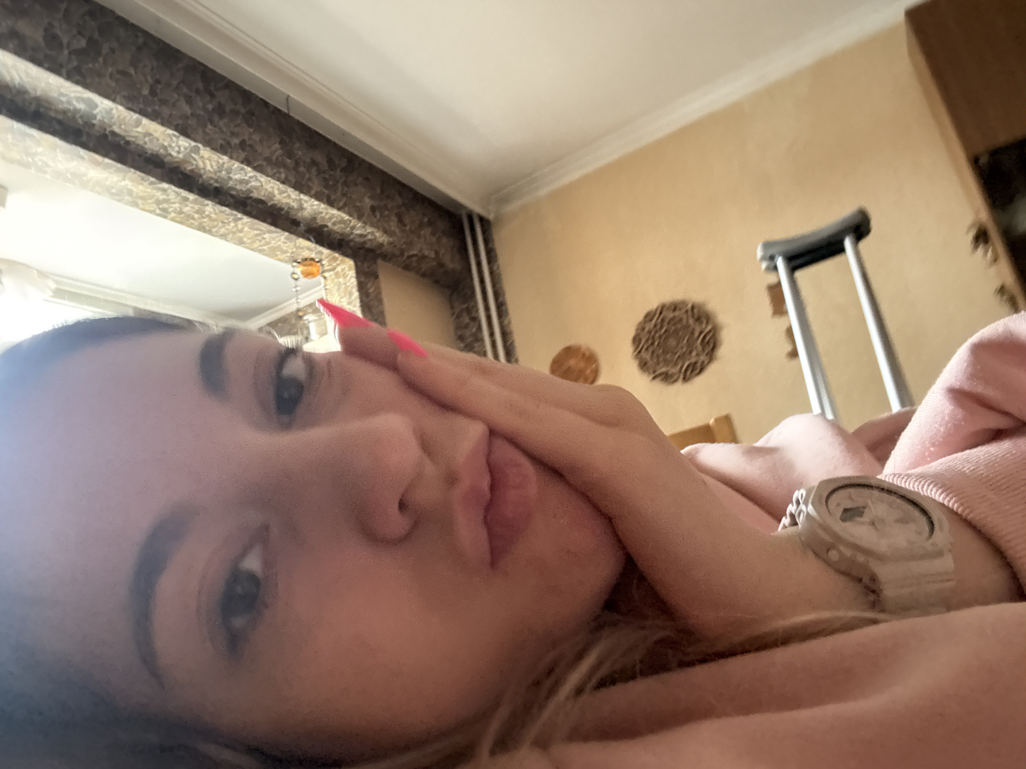 A woman is lying down, likely on a bed, indoors. A crutch is visible in the background. There are decorative items on the wall. The woman is wearing a watch. The location is not discernible as Sofia, Bulgaria based on the image content.