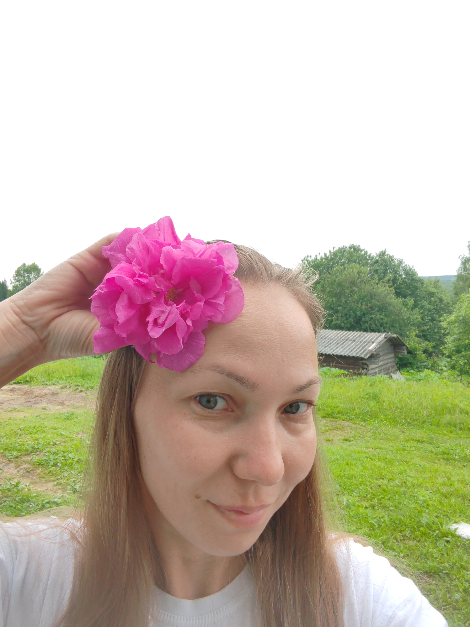 A woman is pictured outdoors with a pink flower on her forehead. She has fair skin, light brown hair, and green eyes. She is wearing a white t-shirt. In the background, there is a wooden building with a corrugated roof, surrounded by lush green grass and trees. The sky is overcast. The location is described as Чухлэм, Russia, suggesting a rural or village setting.