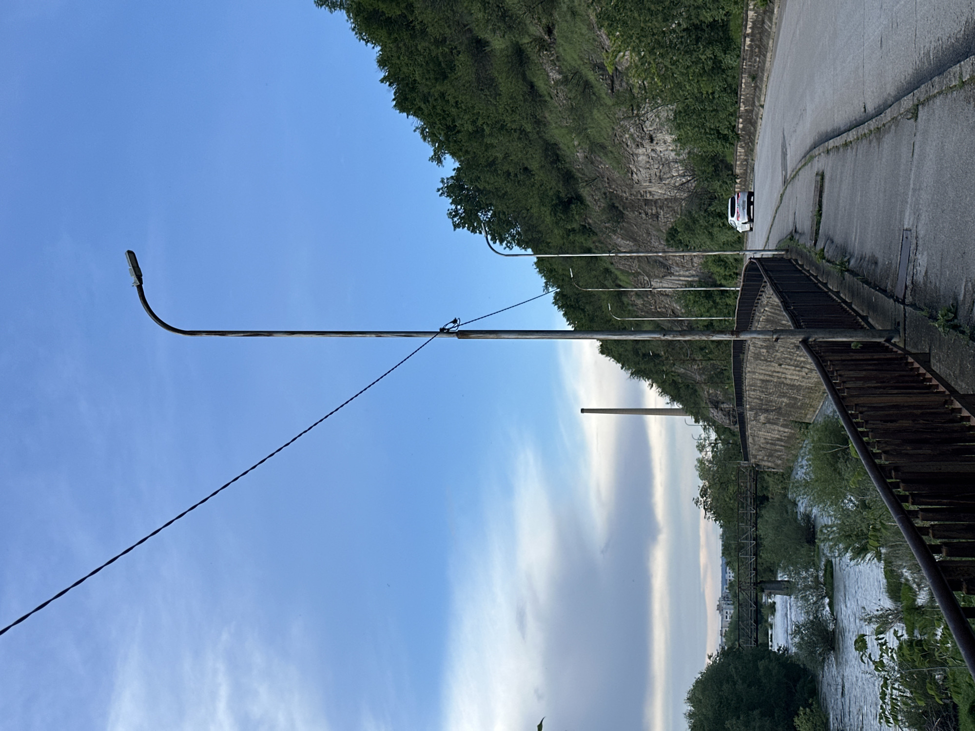 The image shows a view in Lovech, Bulgaria. A paved road curves along a rock face covered with greenery. A white car is parked on the roadside. A metal railing runs along the edge of the road, overlooking a river flowing beneath a bridge. A tall, metal street lamp stands beside the road, with a curved top and a wire extending from its pole. The sky is blue with streaks of clouds.