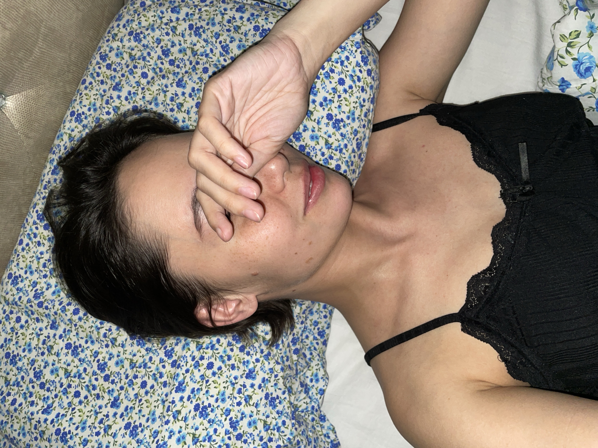 An overhead close-up shot captures a person, likely female, lying on their back on a bed, potentially in Izhevsk, Russia. The individual is wearing a black camisole or sleep top with delicate lace trim along the neckline. Their left hand rests over their face, partially obscuring their eyes and nose, which could suggest sleepiness, waking up, or an attempt to block light. Dark hair is fanned out on a pillow with a distinct blue and white floral pattern. The person's skin shows visible freckles across their face and neck. A white bed sheet covers the mattress, and another pillow with a similar floral pattern is partially visible in the top right. To the left, a textured, possibly upholstered or woven, headboard or wall surface is visible. The scene conveys a sense of quiet relaxation or rest. No text is visible in the image.