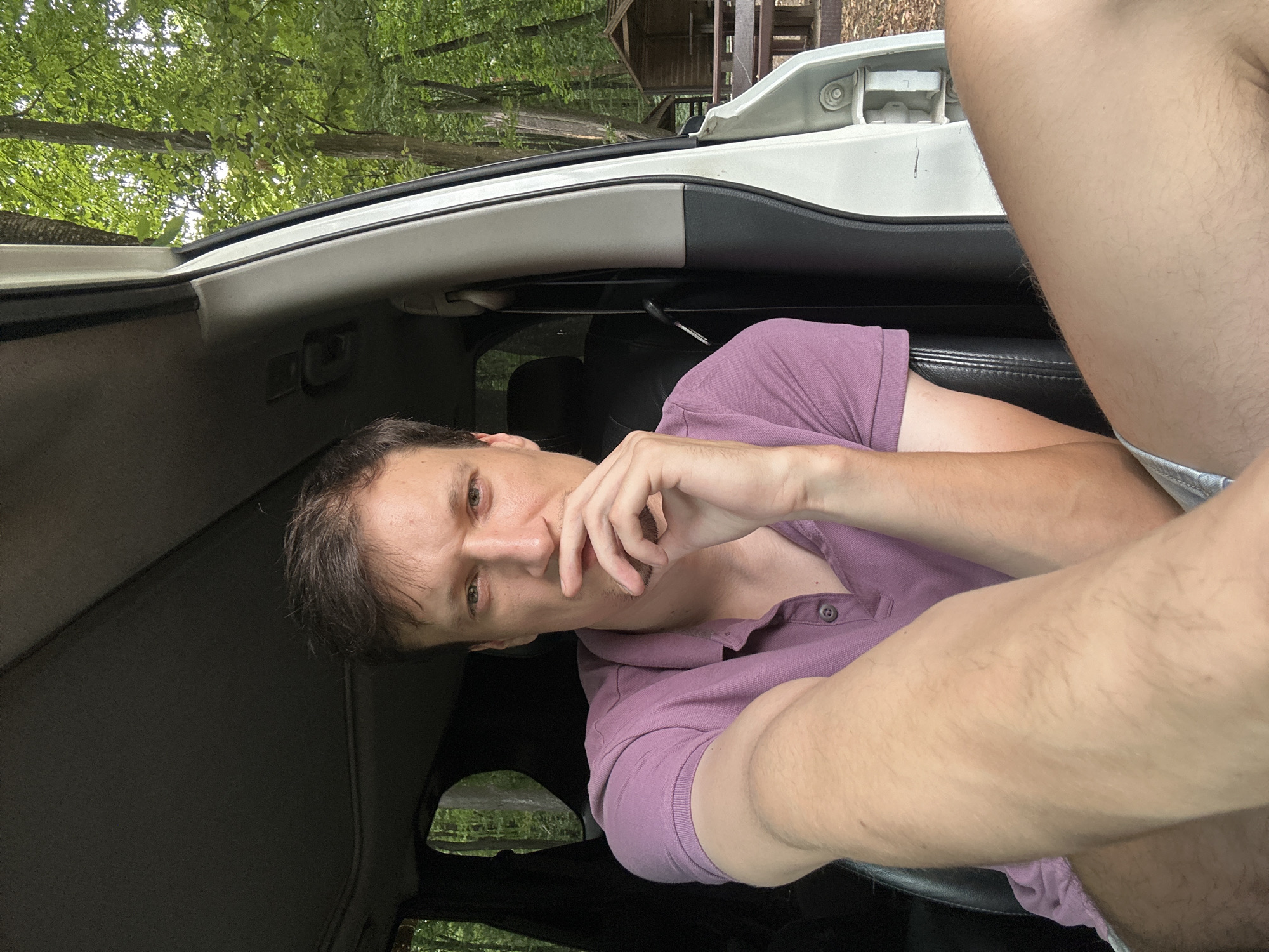 A man with short brown hair and fair skin is seated inside a vehicle, looking towards the camera. He is wearing a purple polo shirt. His right hand is touching his chin, and his left arm is bent. The interior of the vehicle is visible, including the headliner and seats. Through the open door and windows, a wooded area with trees and picnic tables is discernible, suggesting an outdoor or park-like setting. The man's right leg is visible, wearing light-colored shorts.