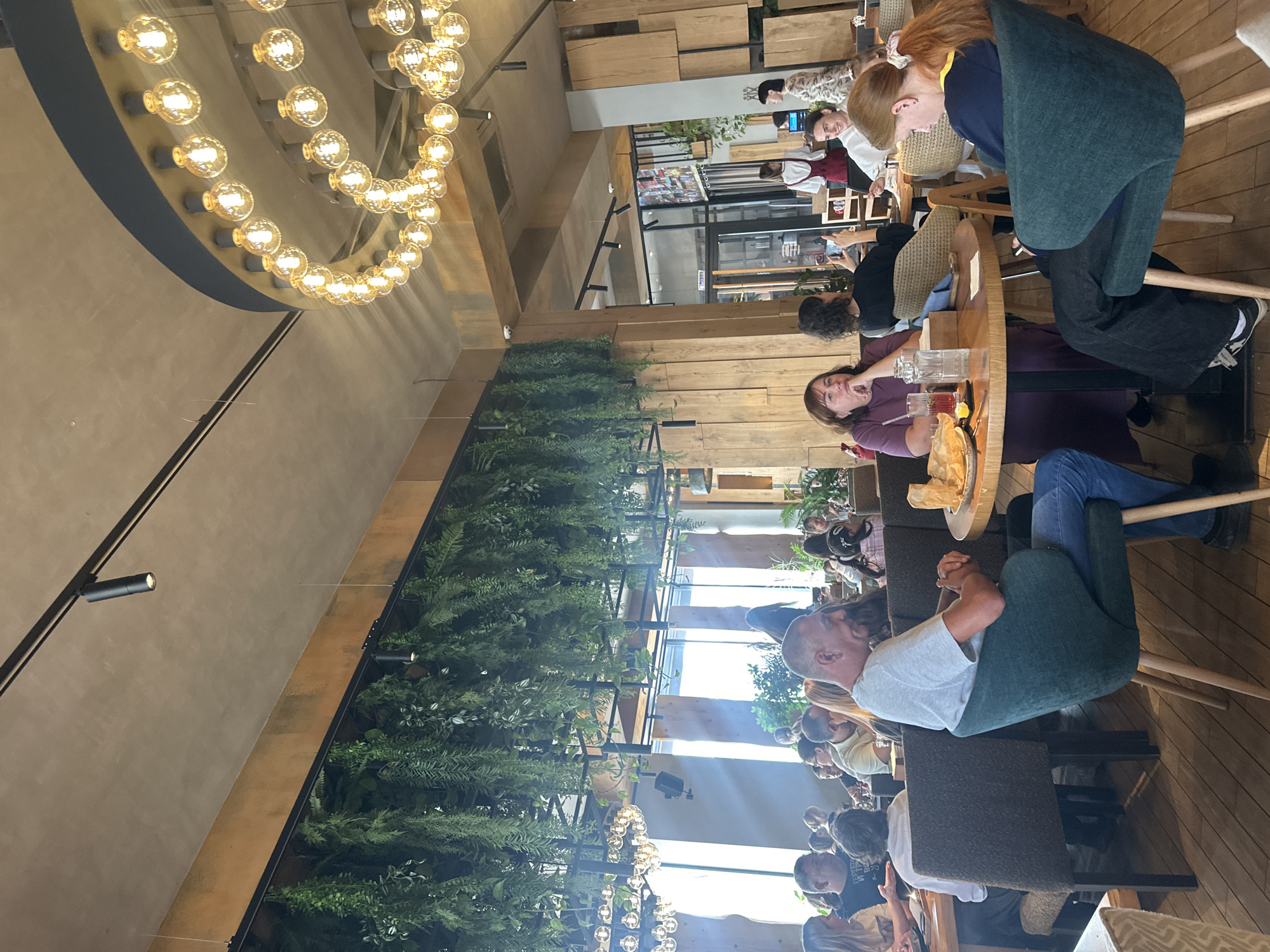 The image depicts the interior of a restaurant with a modern, rustic ambiance. Wooden elements are prevalent in the decor, including the walls, floor, and furniture. Lush green plants hang from the ceiling, adding a natural touch. Several groups of people are seated at tables, engaged in conversations, dining, or using their phones. The lighting is warm and inviting, with a large, circular chandelier featuring numerous Edison bulbs, and track lighting on the ceiling. In the foreground, a man in a grey t-shirt and blue jeans sits in a dark green chair. In the middle ground, a woman in a purple dress is seated at a table with drinks and food. Other diners are visible throughout the restaurant, some interacting, others appearing to be alone. The background shows more seating areas and a glimpse of what appears to be a service area or exit.