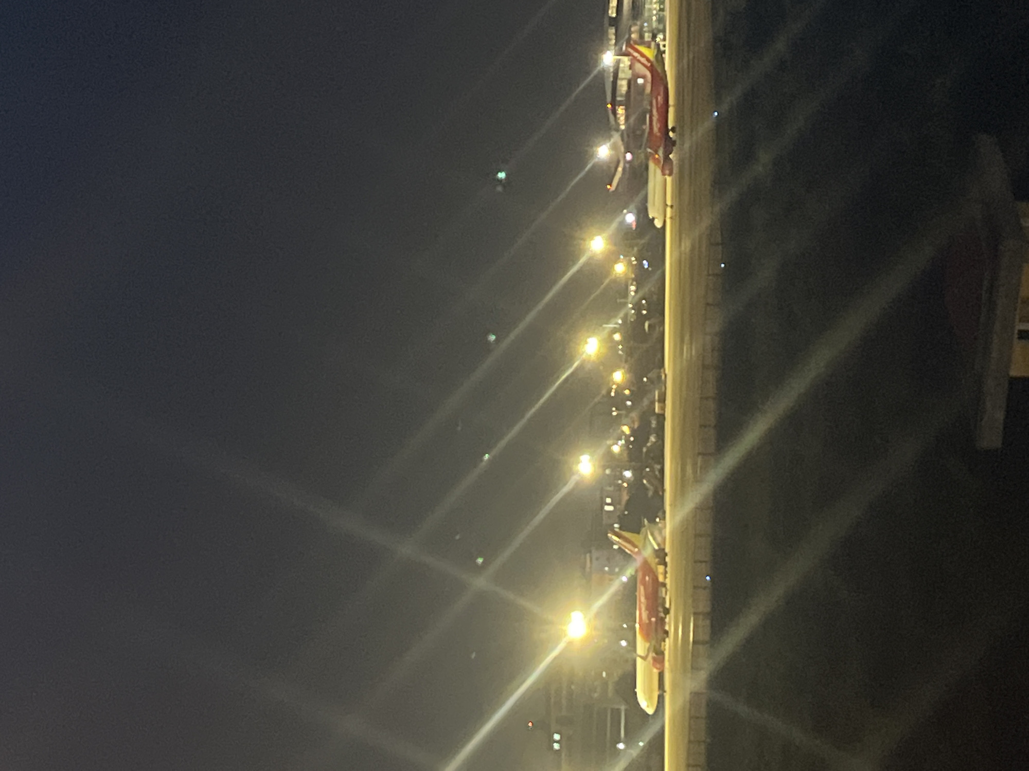 This is a nighttime, eye-level, vertical, outdoor shot of a street scene in Da Nang, Vietnam. The image is dominated by dark, hazy skies and bright, streaking lights, suggesting artificial illumination and possibly some atmospheric moisture.
The main subject in the frame is a section of what appears to be a parade float or a decorated vehicle, depicted on the right side of the image, oriented vertically. It's primarily red and yellow, with some white elements. The float is adorned with lights, and what seem to be people in costumes are visible on it, waving. These figures are difficult to discern in detail due to the lighting and resolution.
Along the left side of the frame, a line of streetlights or decorative lights stretches into the distance, creating prominent light streaks due to the long exposure or lens flare. These lights are a mix of warm yellow and some cooler tones. In the background, numerous small, blurry lights suggest other vehicles, buildings, or possibly spectators. Some green lights are also visible, which might be from aircraft or other illumination sources.
The overall atmosphere is festive and energetic, likely indicating a celebration or event taking place. The heavy darkness suggests it is late at night. The presence of the decorated float and numerous lights points to a public gathering, possibly a festival or a parade. The hazy sky might be due to light pollution or a slightly humid night. There is no visible text in the image.