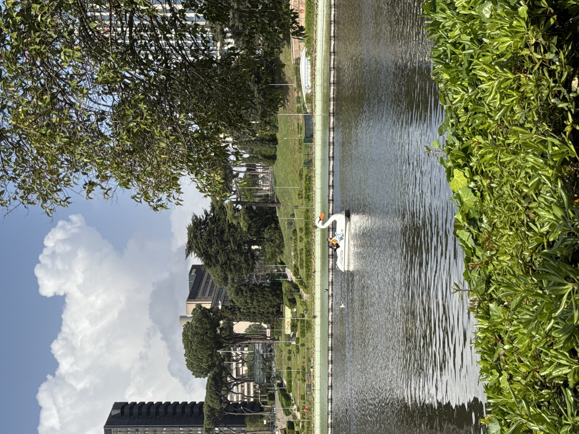 The image depicts a water feature in a park in Rome, Italy. A swan-shaped paddle boat is in the water. Two people are onboard. The water feature is bordered by green foliage and a walkway. Beyond the park are several buildings in an urban setting with a partly cloudy sky.