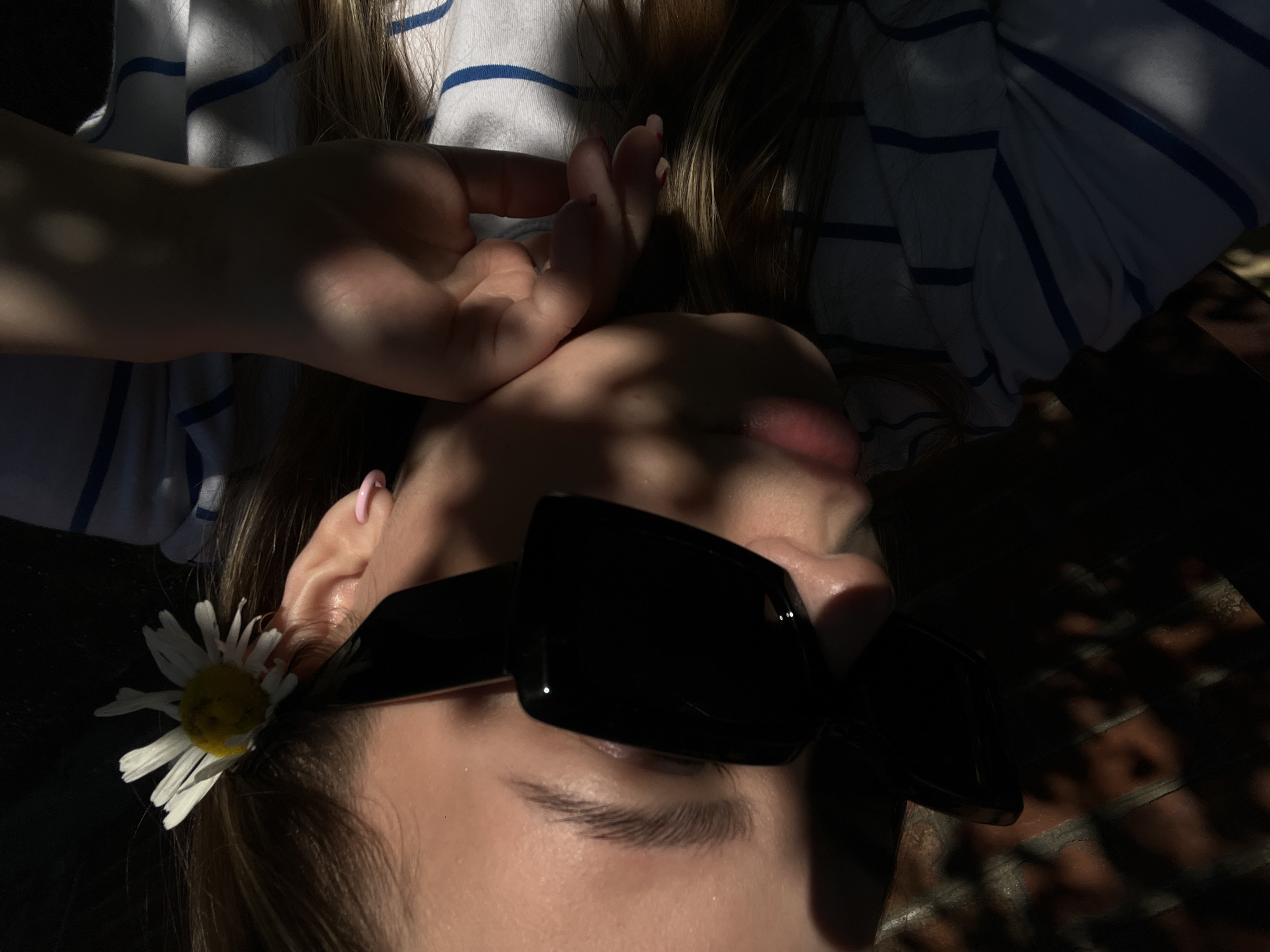 A close-up, overhead shot shows a young woman's face, partially obscured by black sunglasses and dappled sunlight. A white daisy with a yellow center is tucked behind her right ear. She wears a white t-shirt with thin blue stripes. Her left hand rests under her chin, and a small pink hoop earring is visible on her right ear. The background is dark, with the texture of a brick wall visible on the left side, suggesting an outdoor setting. The overall lighting is dramatic, with strong shadows and highlights.