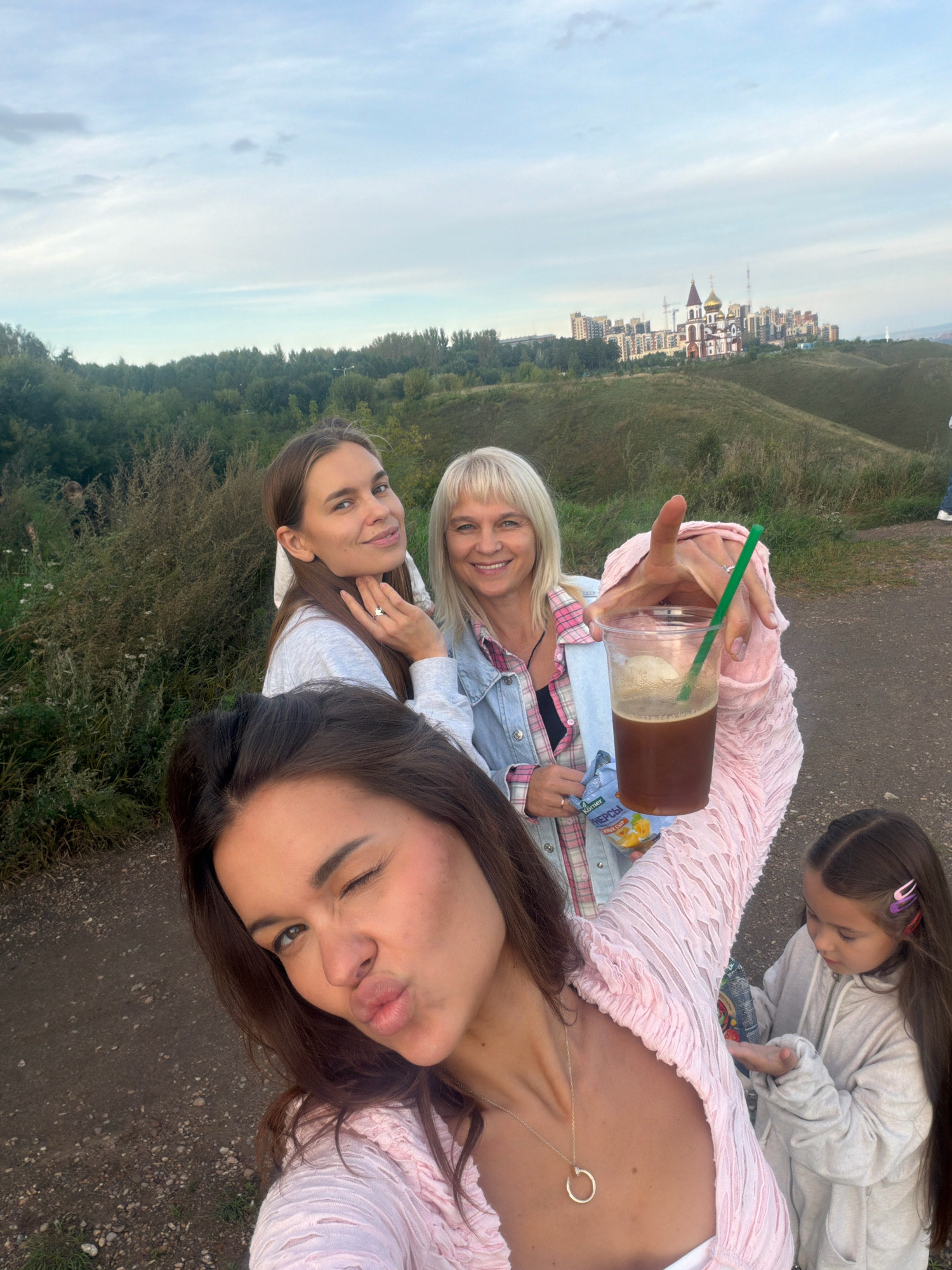 In Krasnoyarsk, Russia, four individuals are captured in a selfie outdoors on a slight incline overlooking a city skyline and a church. In the foreground, a woman with dark hair is pouting her lips, winking, and wearing a pink textured long-sleeved top and a gold necklace. Behind her, a woman with brown hair smiles, wearing a gray sweatshirt and a ring on her left hand. To her right, a blonde woman in a denim jacket over a plaid shirt smiles broadly, holding a clear plastic cup with a brown beverage and a green straw, and a small bag of snacks. To the far right and slightly behind, a young girl with long dark hair and pink and purple hair clips is looking down, wearing a gray hooded sweatshirt. The background features lush green trees, rolling hills, and a prominent church with golden domes alongside modern apartment buildings. The sky is overcast with soft clouds.